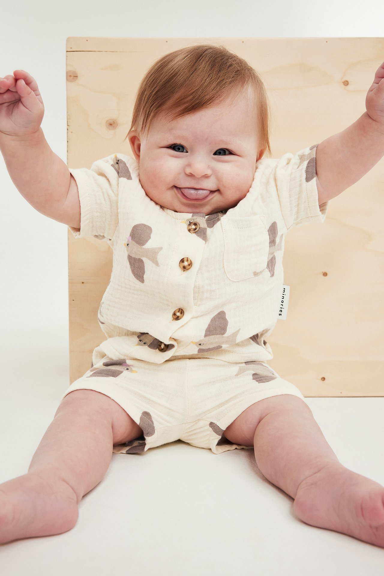 Framifrån, glad baby i beige djurmönstrad muslinkvalitet, tvådelat set med kortärmad skjorta och shorts.