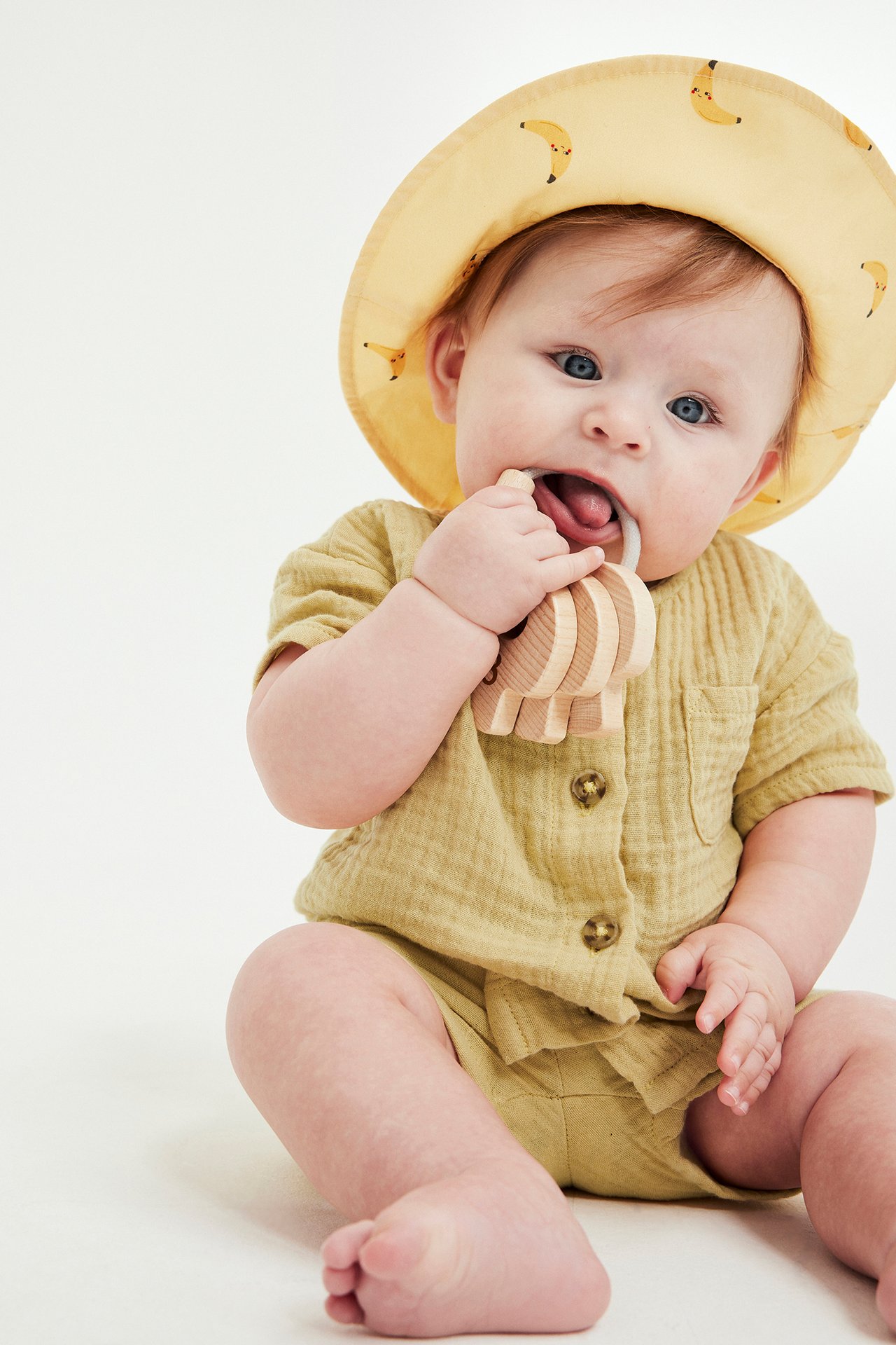 Från framsidan: baby i gult muslinset, kortärmad skjorta och shorts, bananhatt, håller trätandleksak.