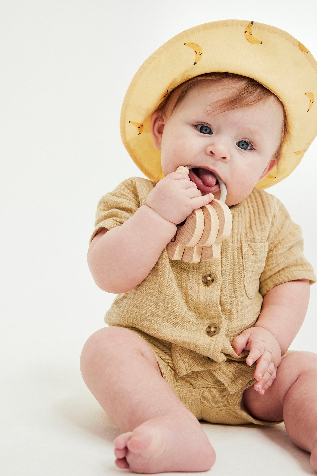 Vauva keltaisessa musliinipaidassa ja shortseissa, keltaisessa banaanihatussa, pureskelee puista purulelua. Edestä.