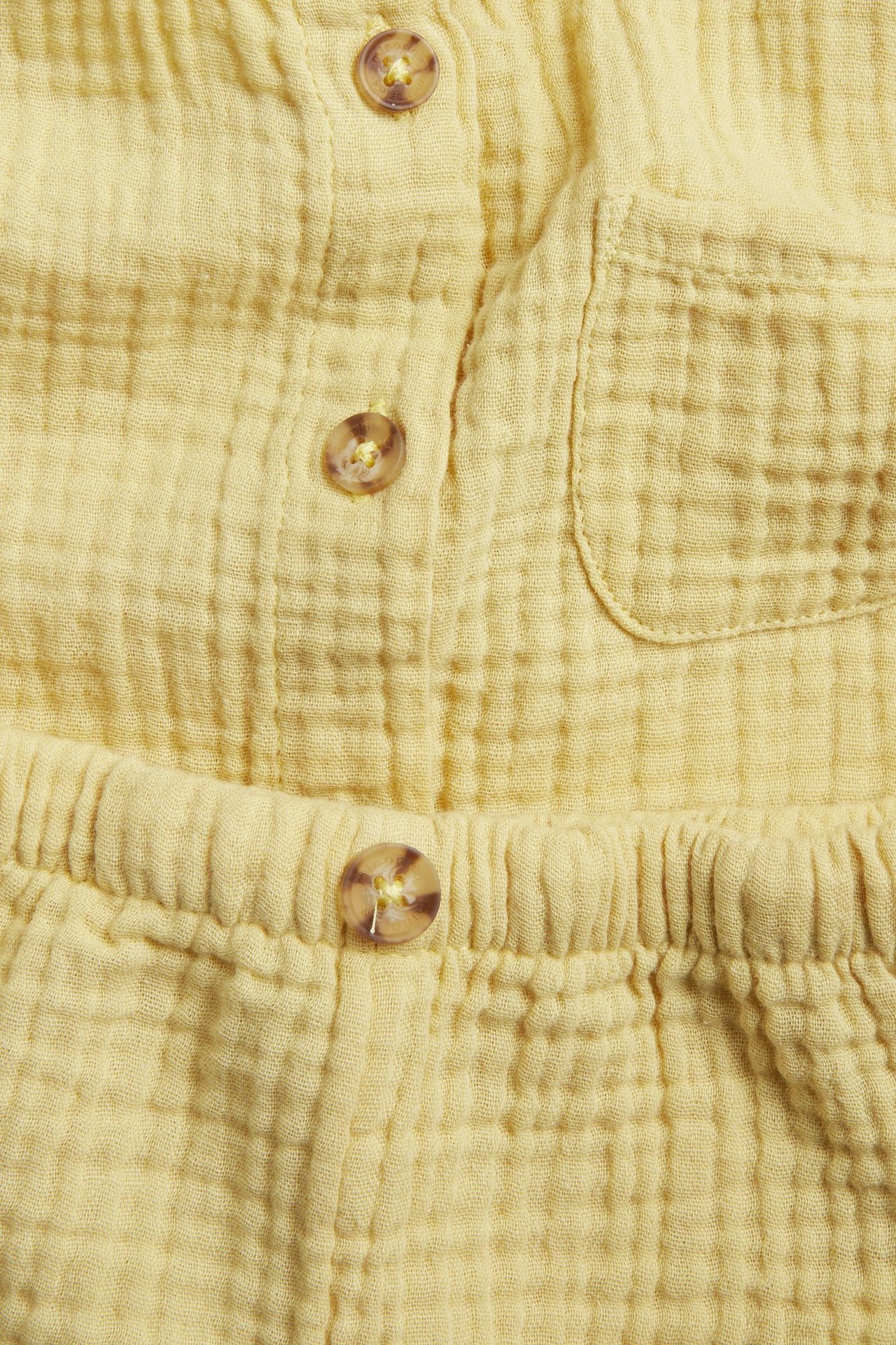 Närbild framifrån på ett gult enfärgat babyset i muslin: skjorta med knappar, ficka och shorts med resår i midjan.