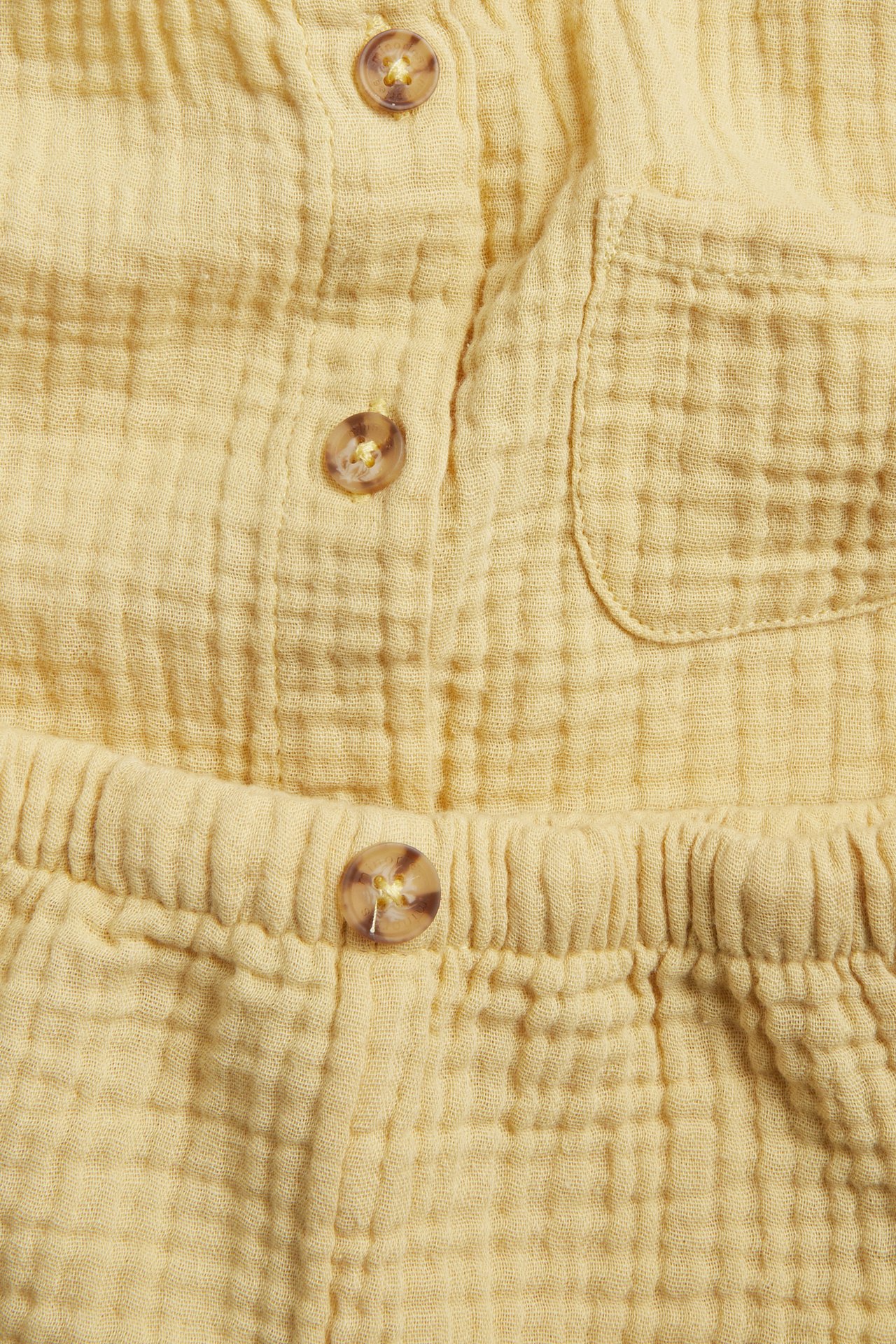 Zbliżenie na jasnożółty niemowlęcy komplet muślinowy: koszula z guzikami, krótkim rękawem i szorty z gumką.