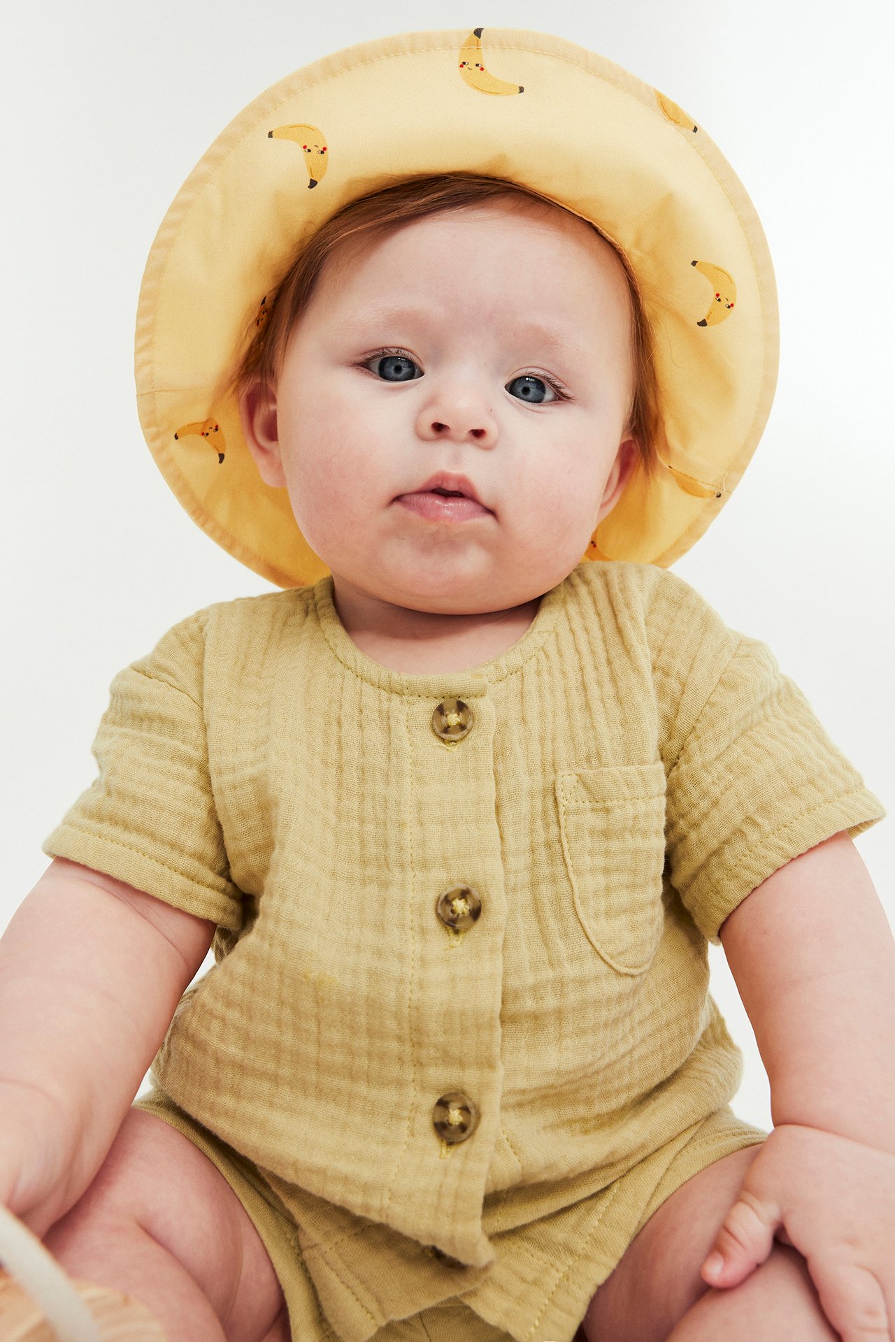 Frontvy av en baby i gult kortärmat muslinsset med skjorta, shorts och en hatt med bananmönster.