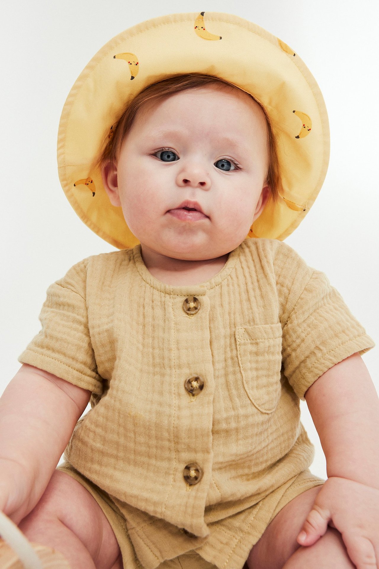 Edestä katsottuna, vauva keltaisessa musliinipaita- ja shortsiasussa, päässä keltainen banaanikuvioinen hattu.