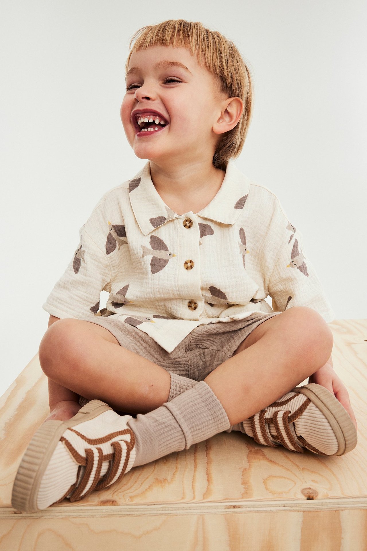 Hymyilevä lapsi beigessä lyhythihaisessa musliinipaidassa lintukuvioilla, vaaleissa shortseissa ja beigenvärisissä tennareissa.