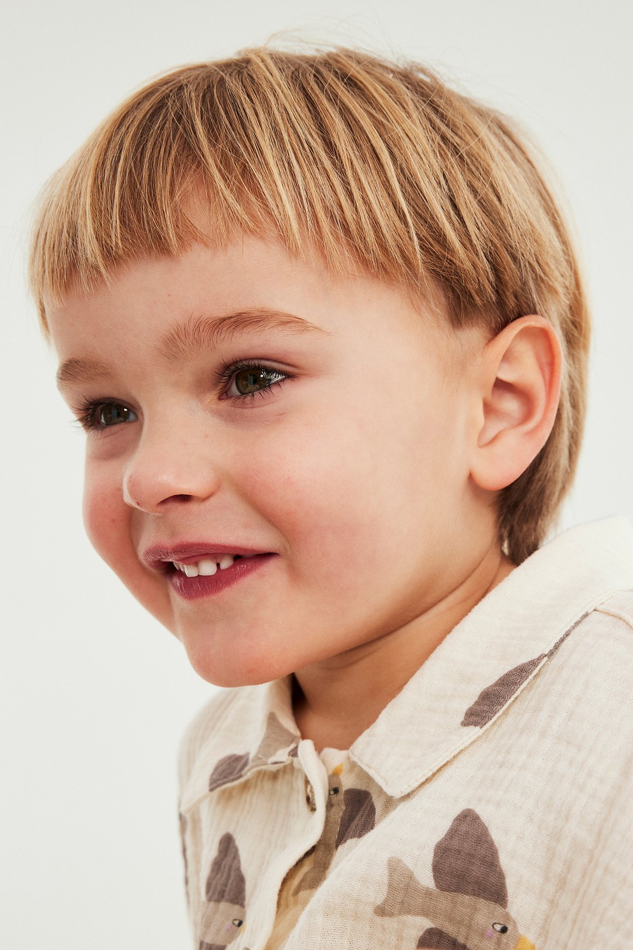 Lähikuva hymyilevästä lapsesta beigessä kuviollisessa lyhythihaisessa musliinipaidassa kauluksella.