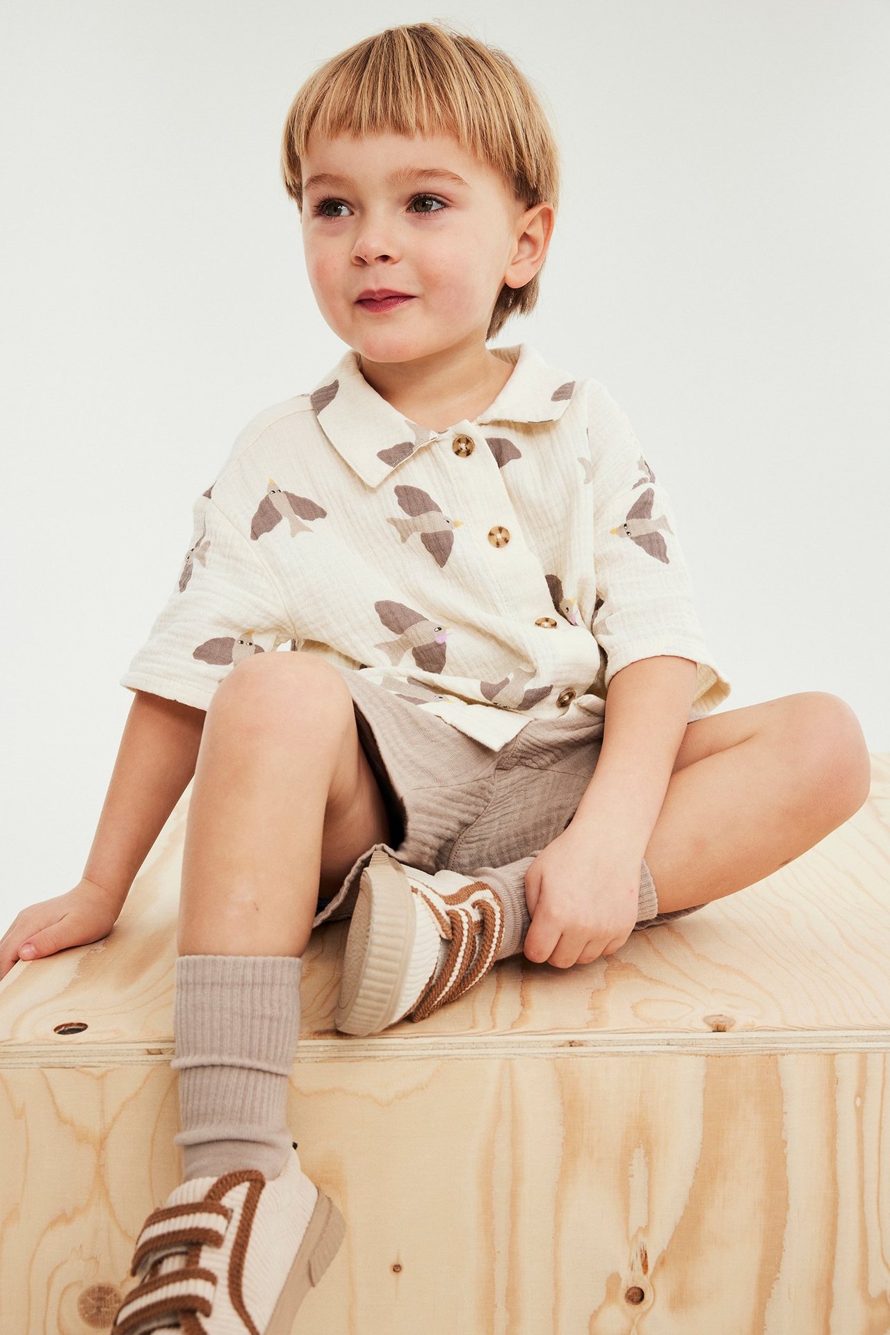 Edestäpäin, lapsi yllään beige kuviollinen lyhythihainen musliinipaita, vaaleanruskeat shortsit, beiget ribbisukat ja tennarit.