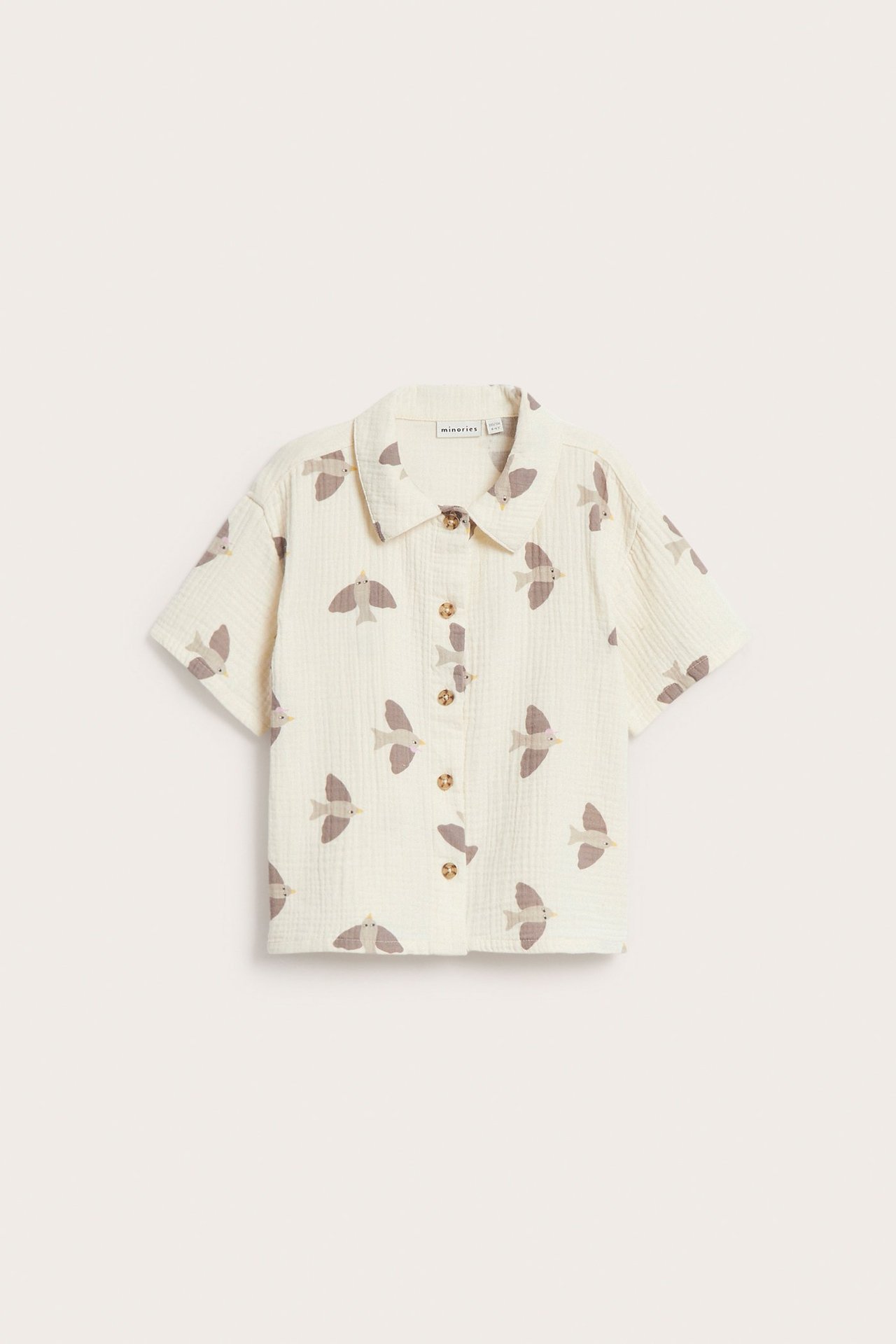 Edestä, vaaleanbeige lasten lyhythihainen musliinipaita ruskeilla lintukuvioilla ja napeilla edessä.