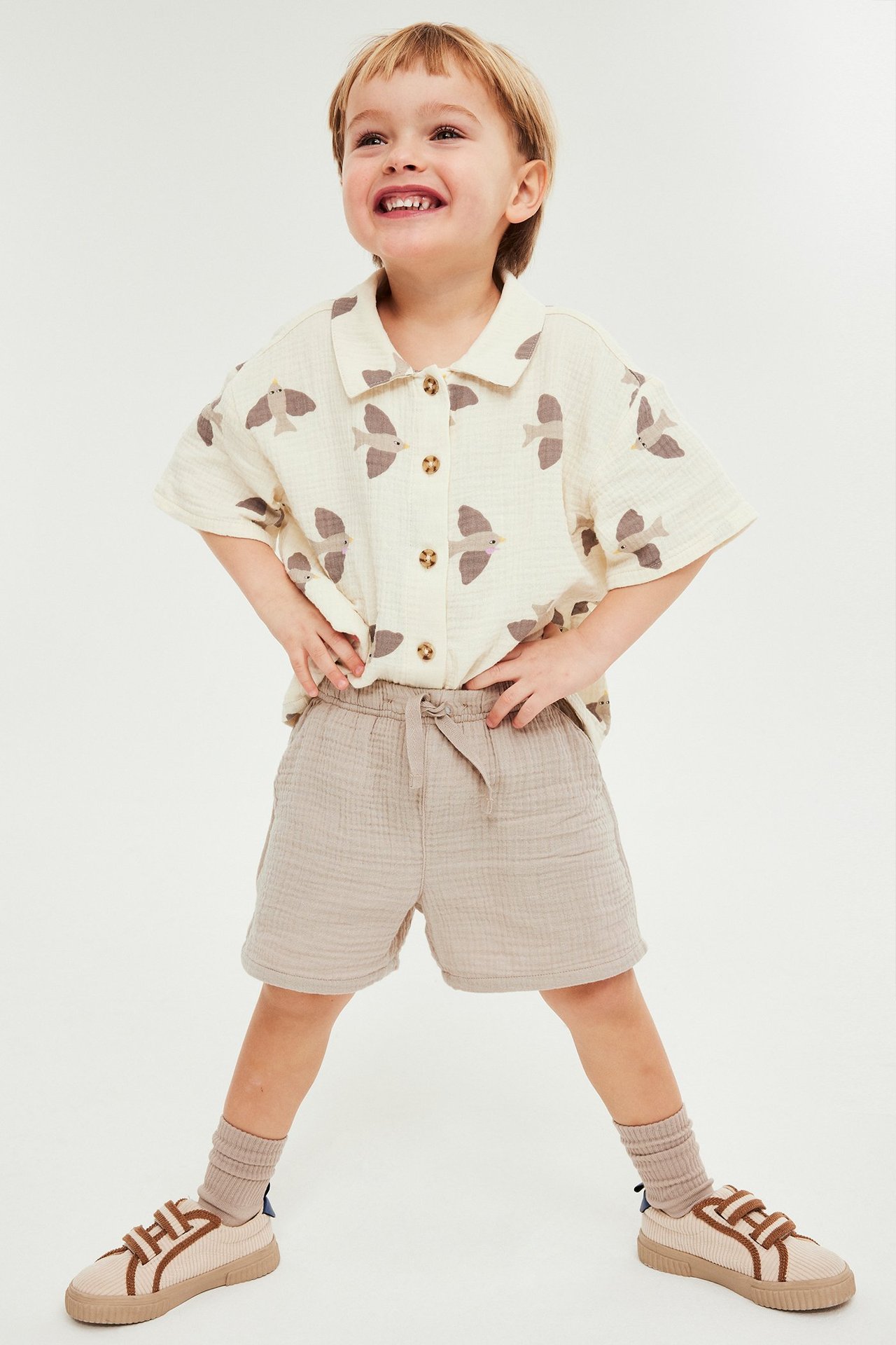 Frontbilde av smilende barn i beige musselinshorts, en beige fuglemønstret skjorte, sokker og joggesko.