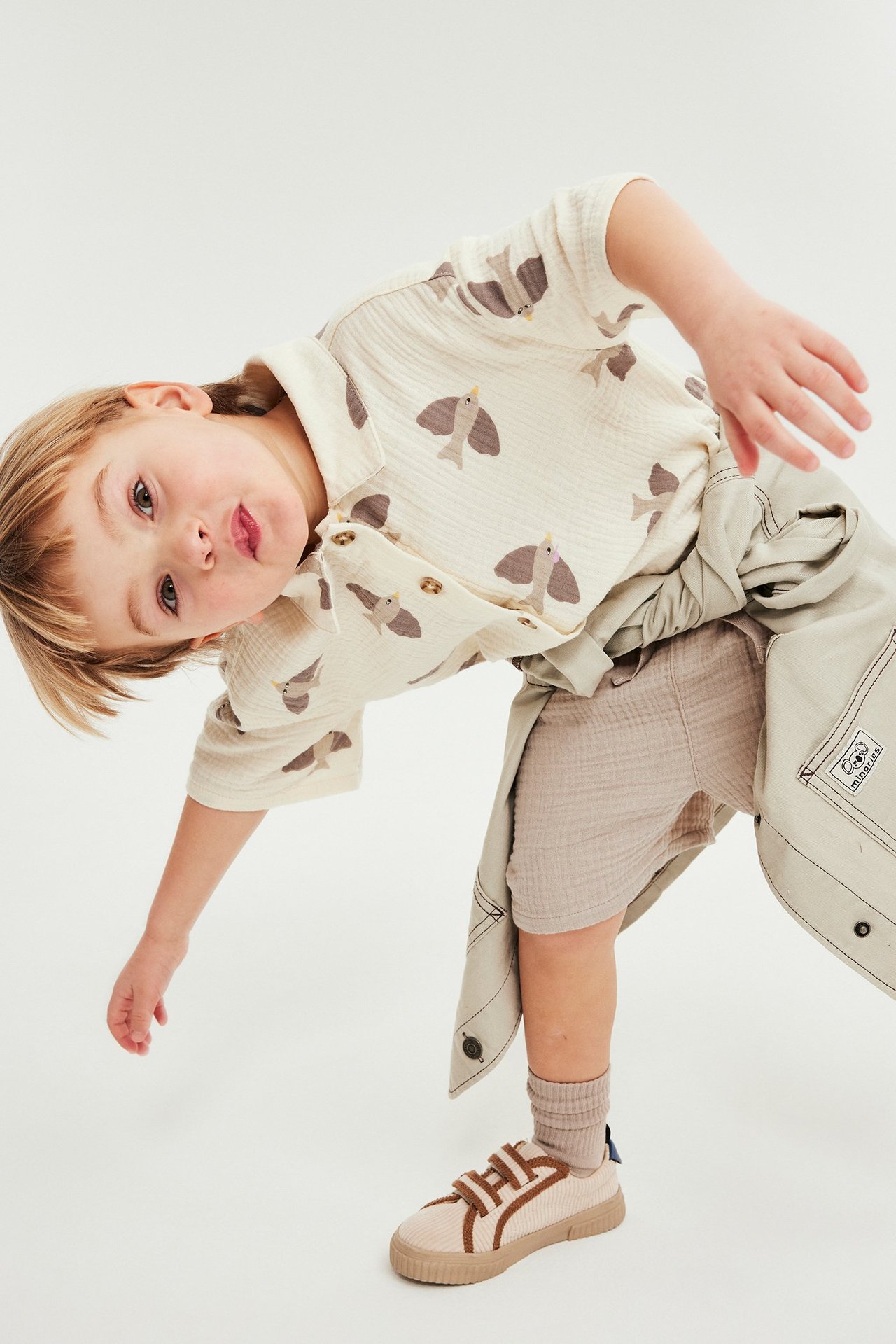 Forfra: Barn i beige muslinsorts, off-white fuglemønstret skjorte og beige joggesko. En jakke er knyttet i midjen.
