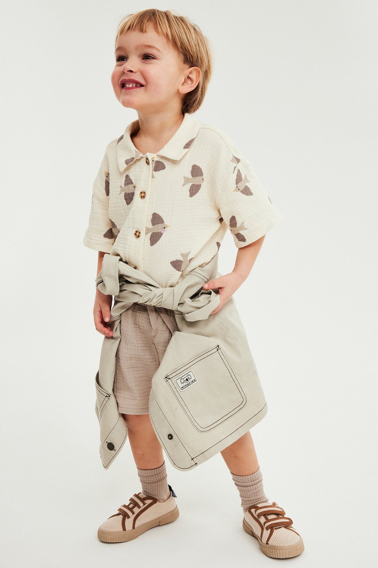 Forfra: Barn i beige muslinshorts, kremfarget fugletrykt skjorte, knyttet beige jakke, beige sokker og borrelåssko.