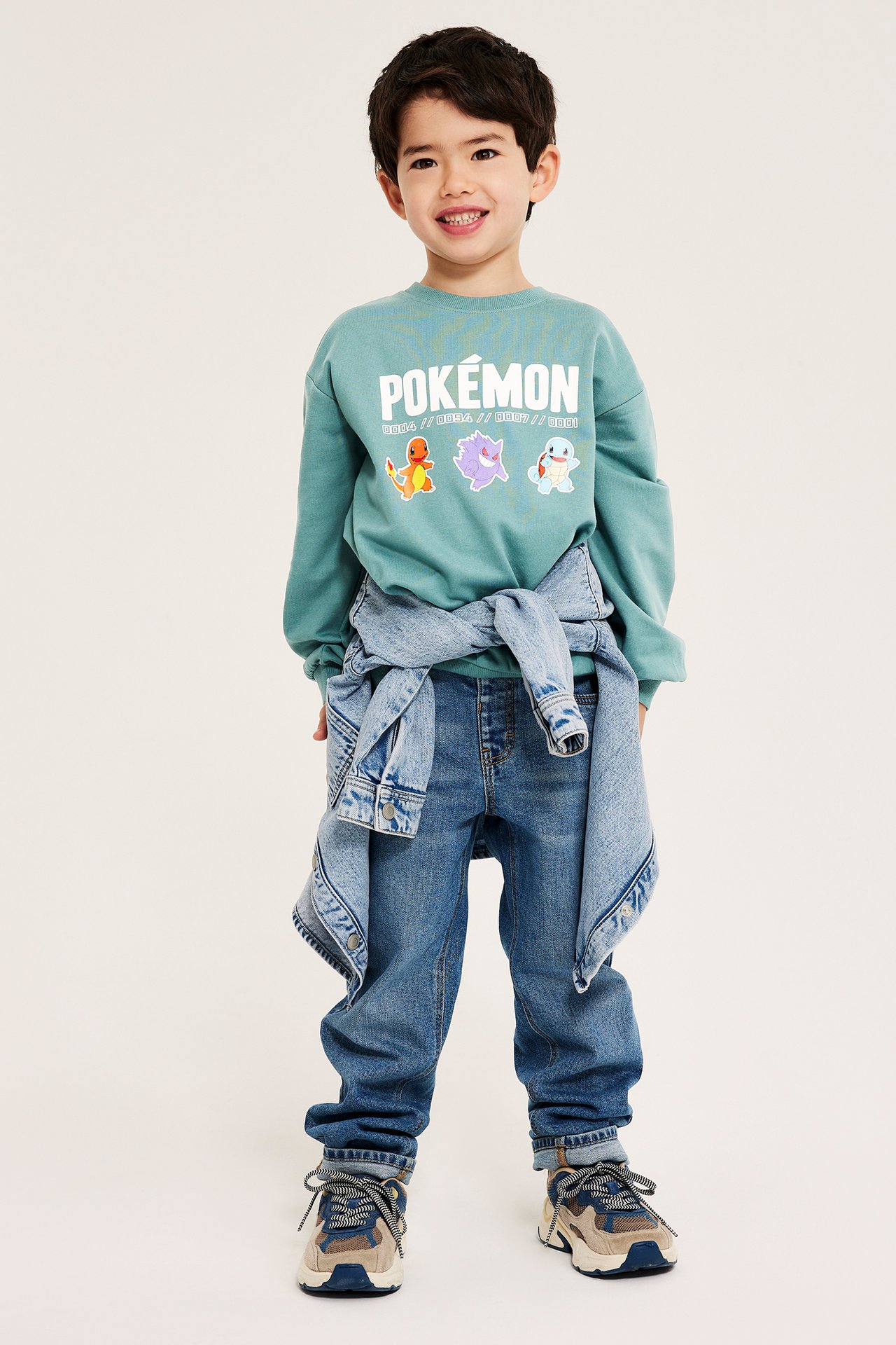 Framifrån: Barn i ljusgrön bomulls-Pokémon-sweatshirt, blå jeans, denimjacka i midjan och sneakers.