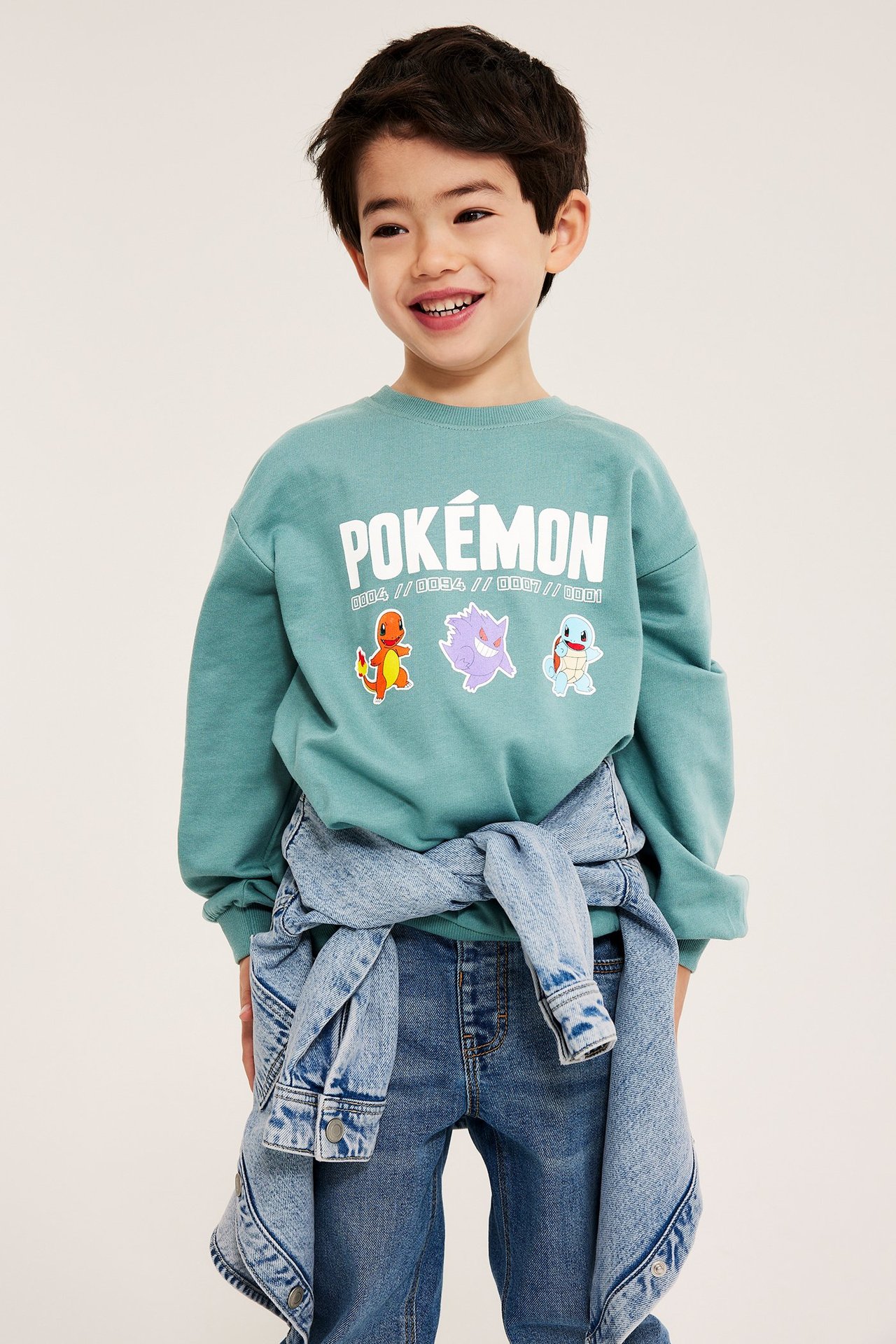 Widok z przodu: dziecko w jasnozielonej bluzie Pokémon, niebieskich dżinsach, związanej kurtce dżinsowej.
