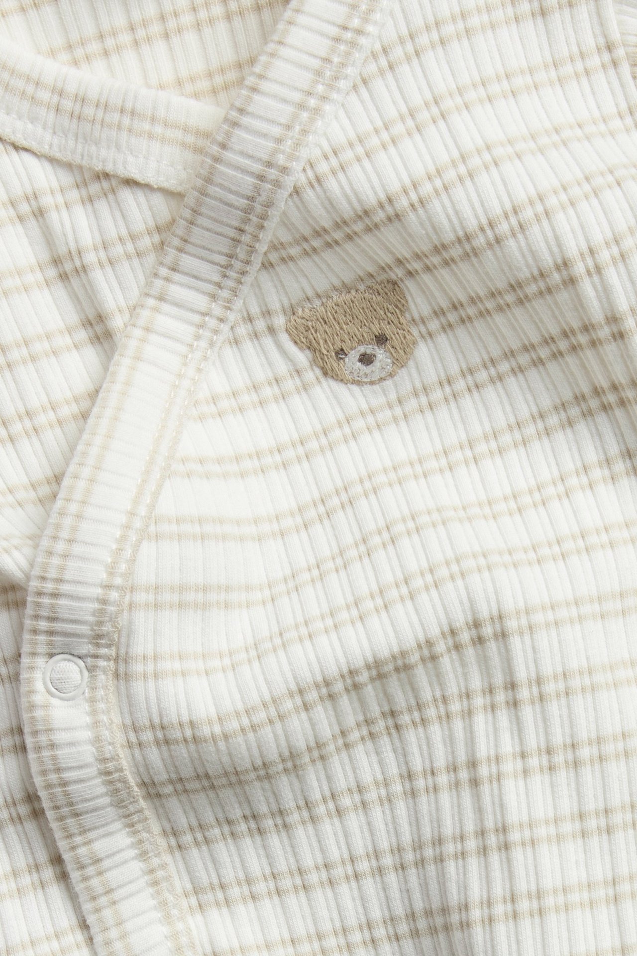 Nærbilde av en beige stripete ribbet baby-omslagstbody med en liten brodert bamse på brystet.