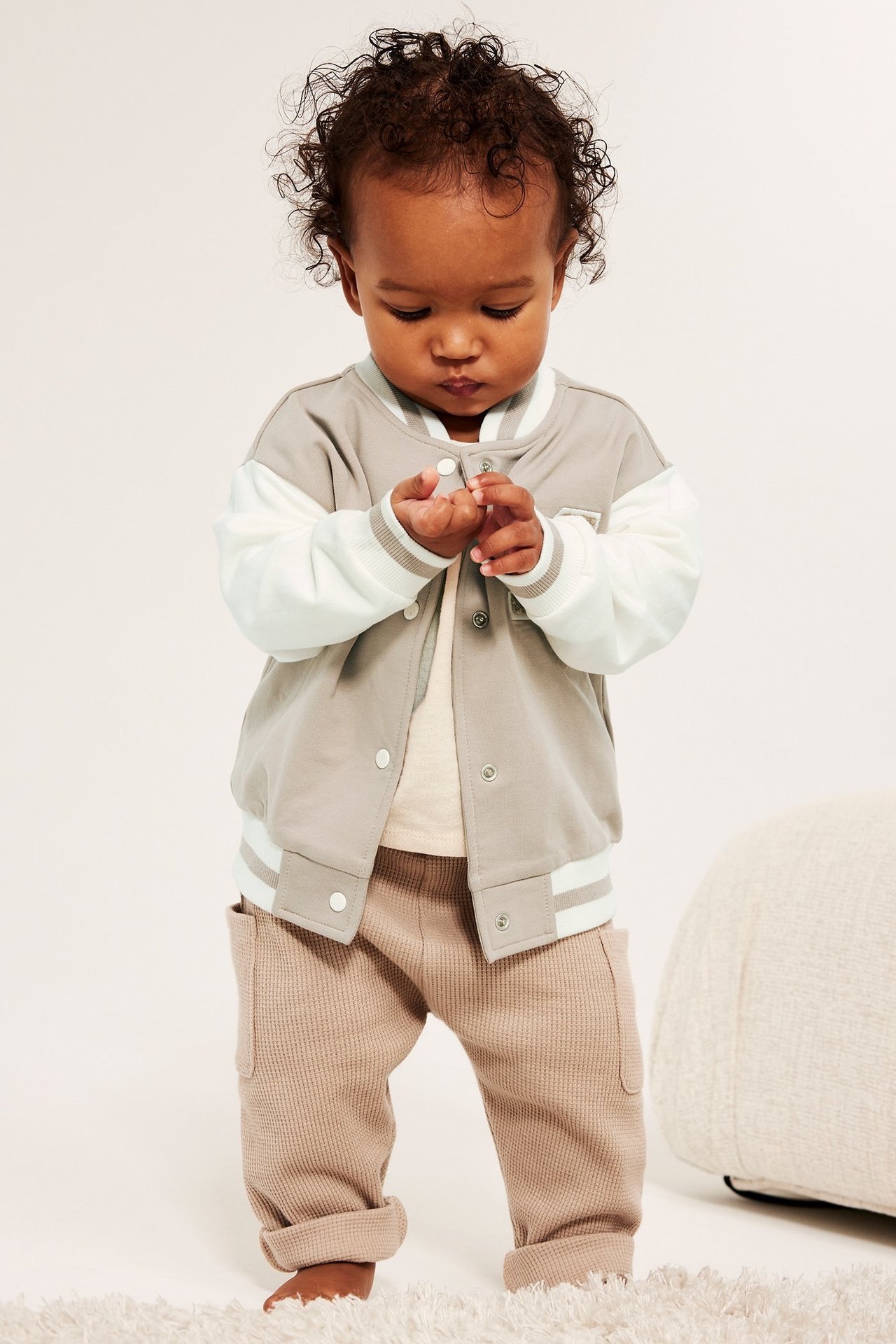 Frontvisning av baby i beige og hvit baseballjakke med trykknapper, over beige t-skjorte og vaffelbukser.