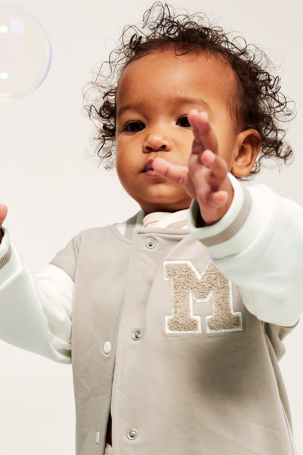Frontbilde: baby i en beige baseballjakke med hvite ermer, trykknapper og 'M'-applikasjon.