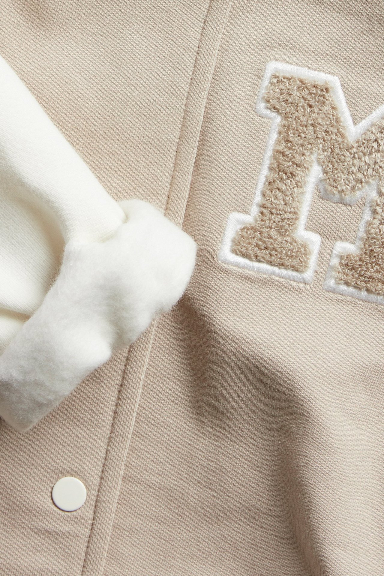 Nærbilde av en beige bomullsblanding baseballjakke med hvit trykknapp, brettet mansjett og frotté M-applikasjon.