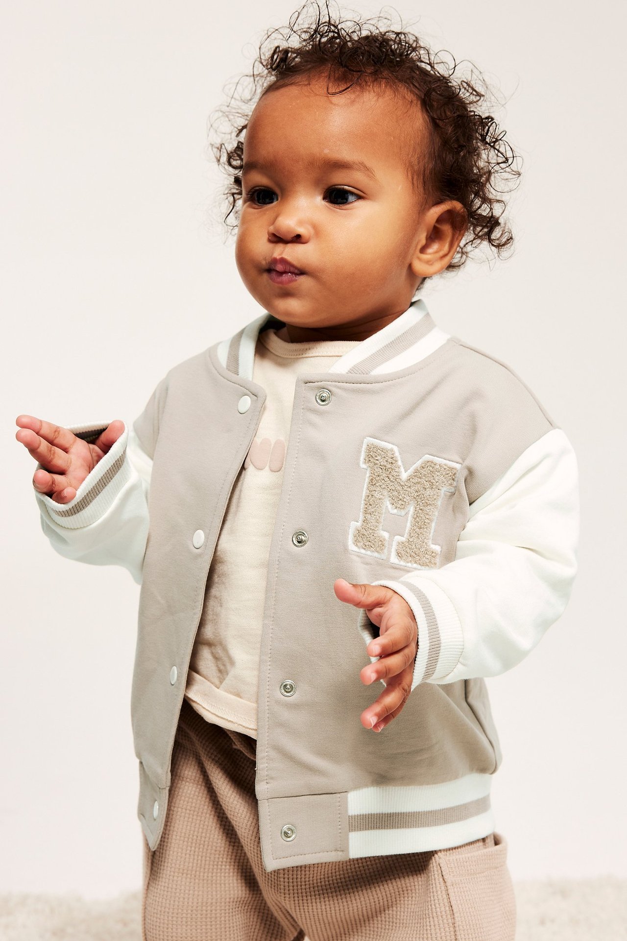 Baby har på seg en beige og hvit baseballjakke med 'M'-applikasjon, over en lys beige topp.