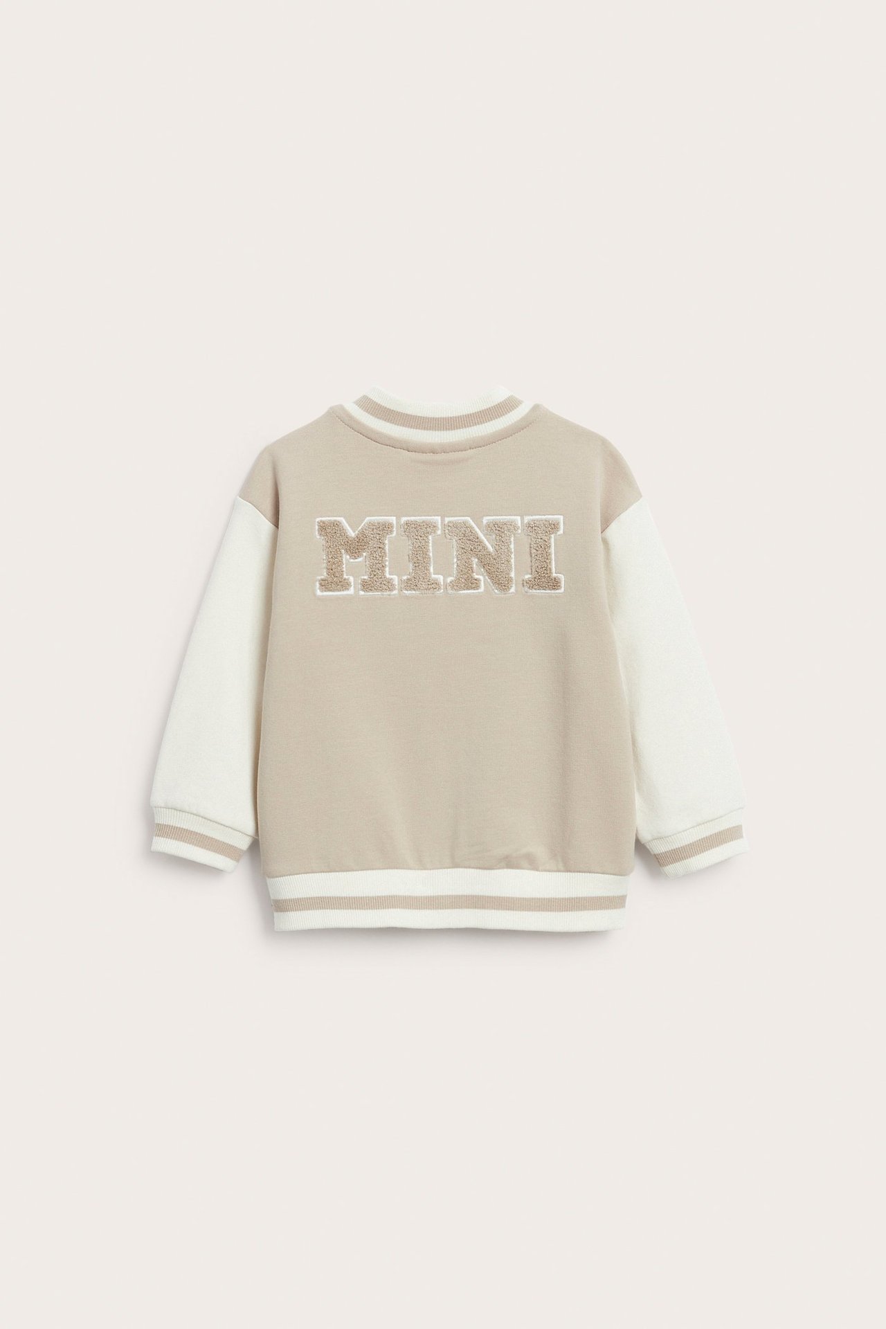 Frontbilde av en beige og off-white baseballjakke for baby med 'MINI' frottéapplikasjon på brystet.