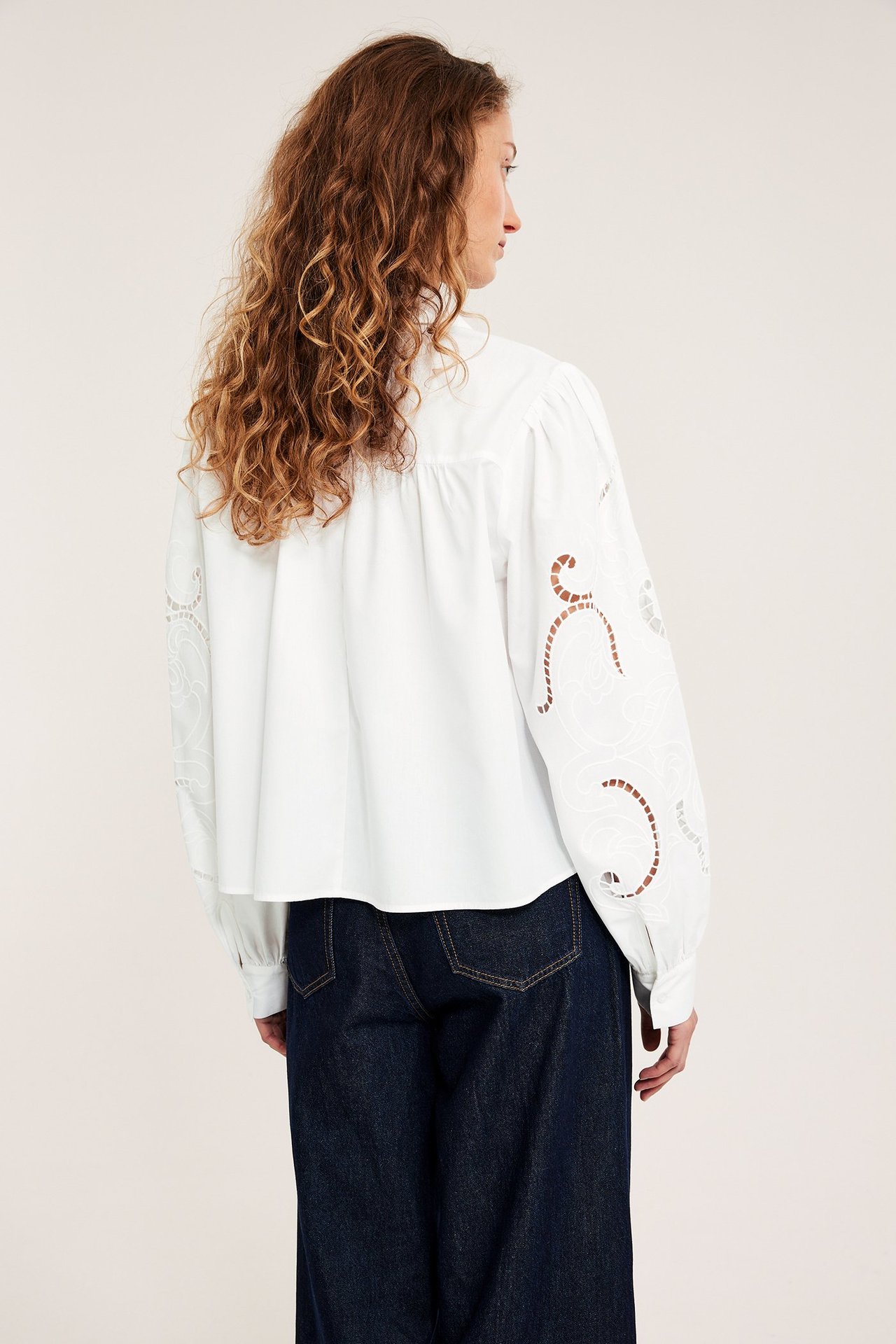 Rygg-/sidebilde av kvinne i hvit, kort bomullsbluse med broderte ermer og mørkeblå jeans.