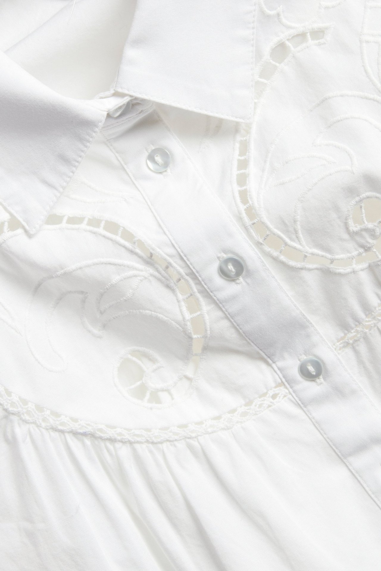 Nærbilde av en hvit bomullsbluse med brodert knappestolpe, med utskårne mønstre og lyse knapper.