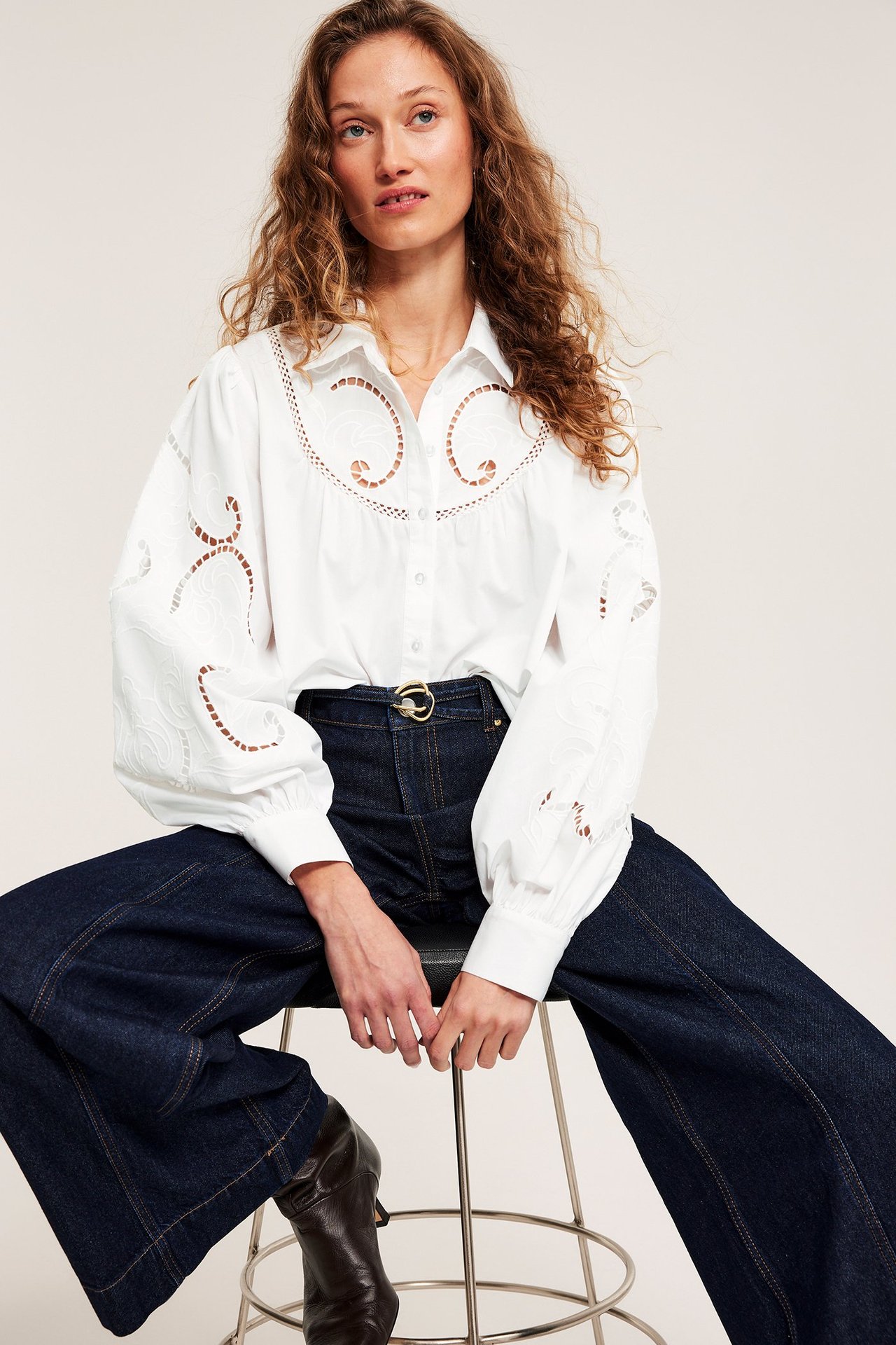 Front: Kvinne i hvit brodert bomullsbluse, mørkeblå jeans, gullbelte og brune støvler.