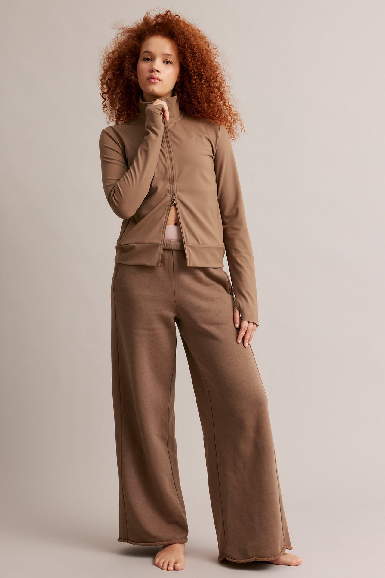 Framifrån: Kvinna i brun åtsittande träningsjacka med hel dragkedja, tumhål och matchande bruna vidbenta mjukisbyxor.