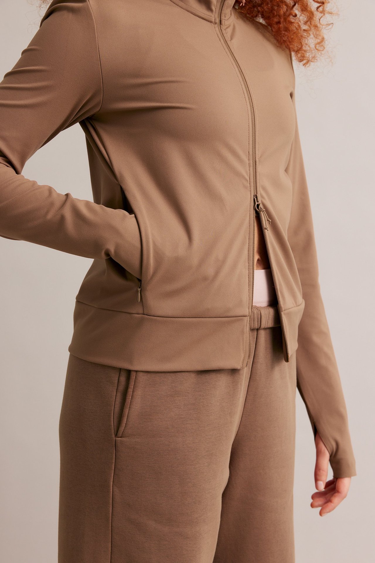 Kvinna i brun träningsjacka med dragkedja, tumhål, fickor och matchande bruna byxor.