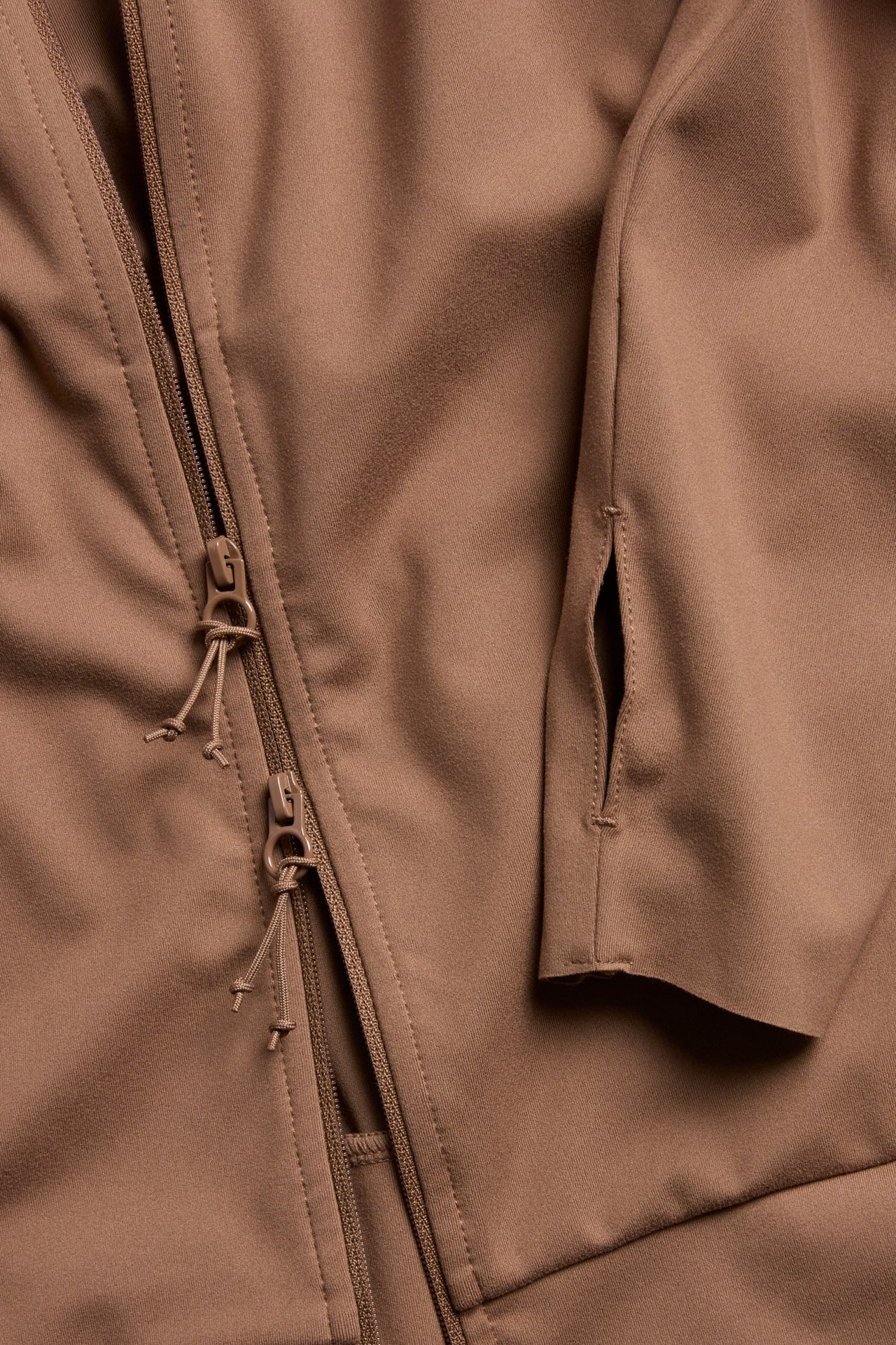 Zbliżenie na brązową kurtkę treningową z zamkiem błyskawicznym i otworem na kciuk w rękawie.