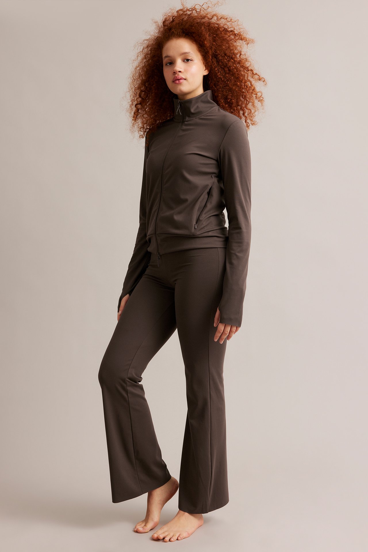 Framifrån: kvinna i brun träningsjacka med dragkedja, tumhål och matchande utsvängda byxor.