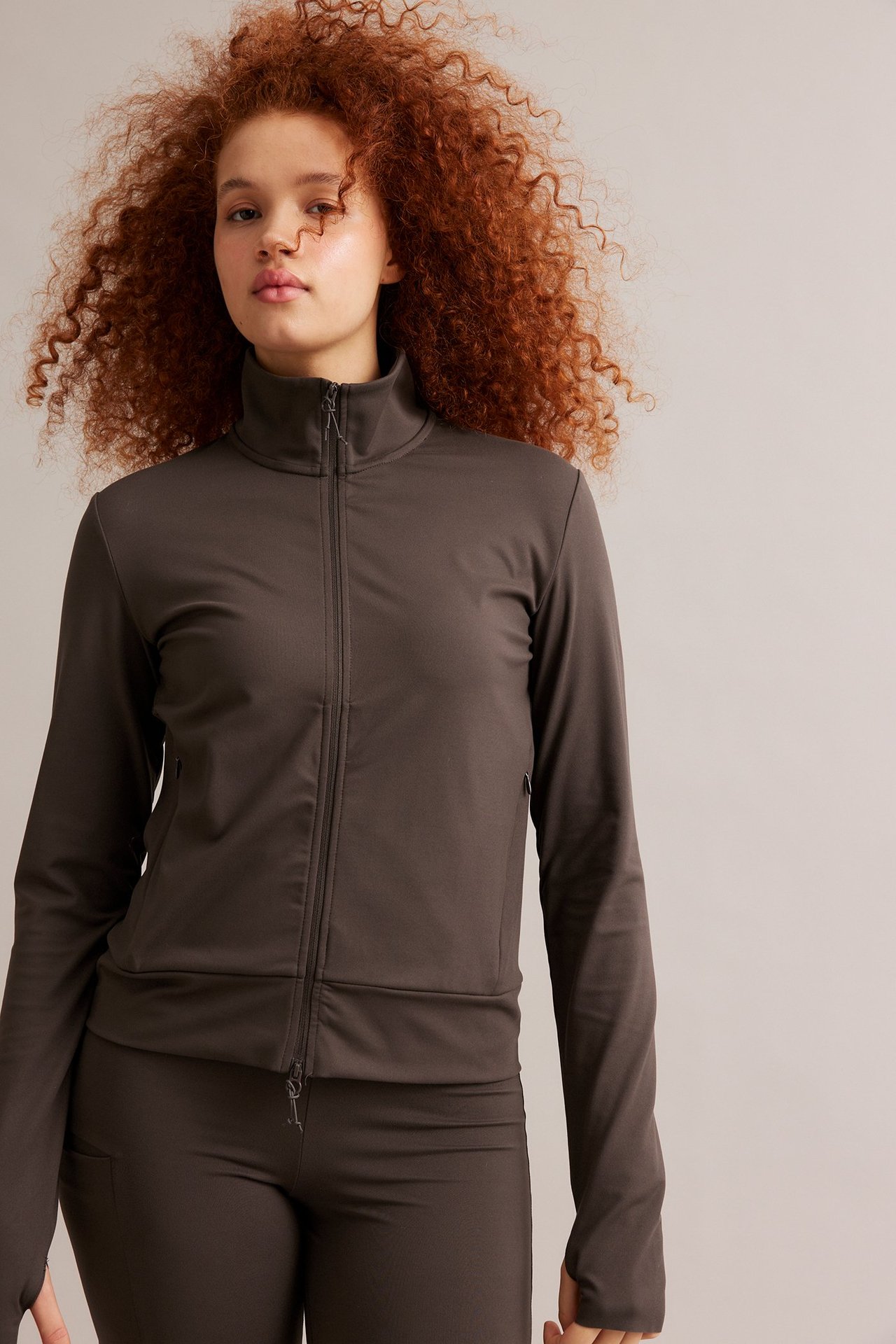 Framifrån: kvinna i brun träningsjacka med hel dragkedja, tumhål, fickor och matchande byxor.