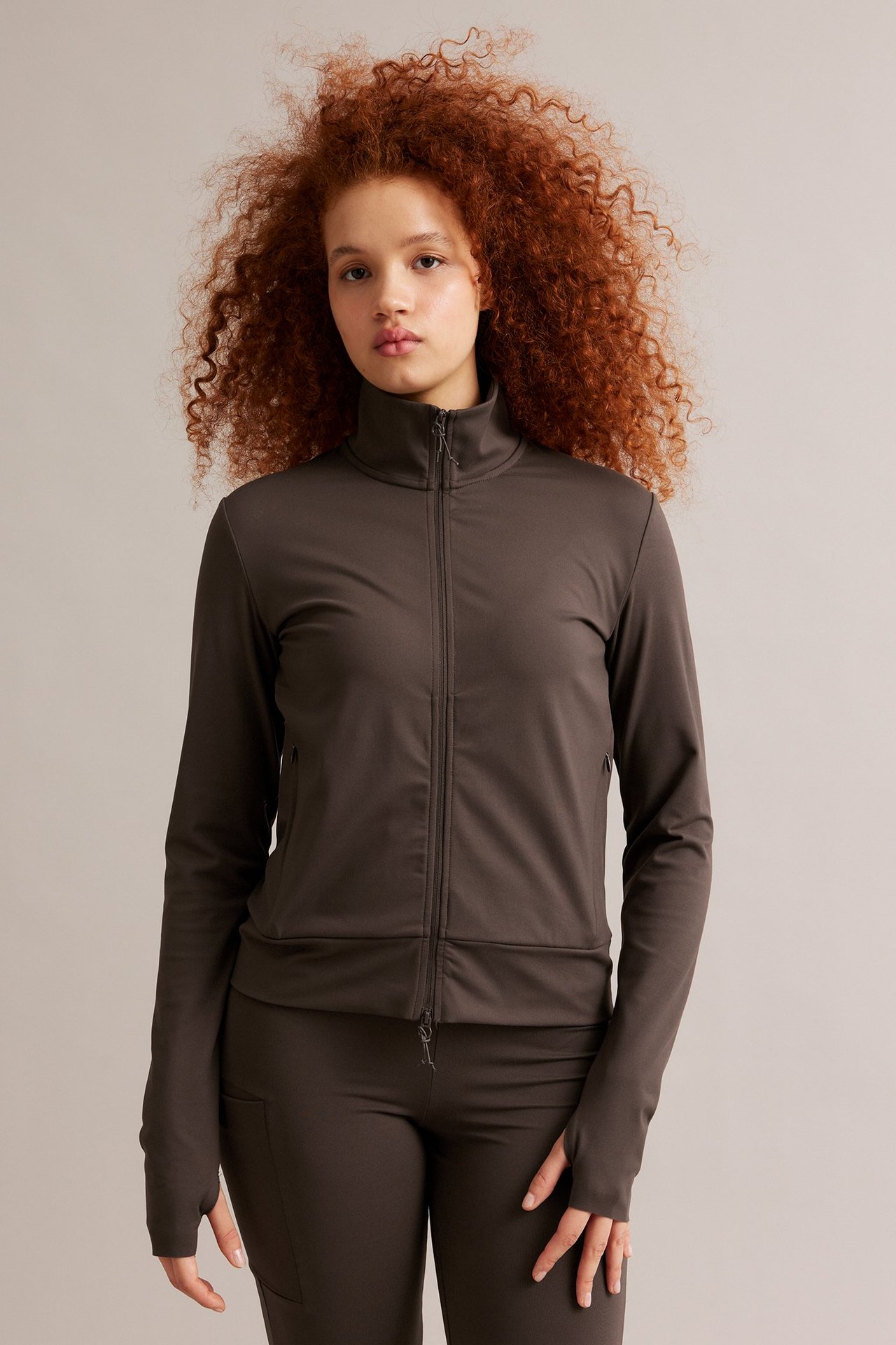Frontvy av kvinna i brun träningsjacka med hel dragkedja, tumhål och matchande byxor.