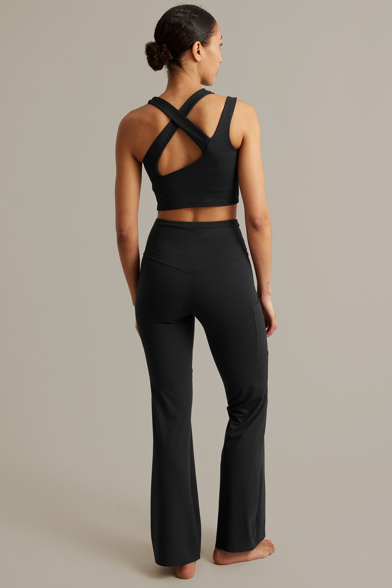 Naisen takakuva: mustat korkeavyötäröiset bootcut-treenileggingsit sivutaskulla ja musta ristikkäisselkäinen toppi.