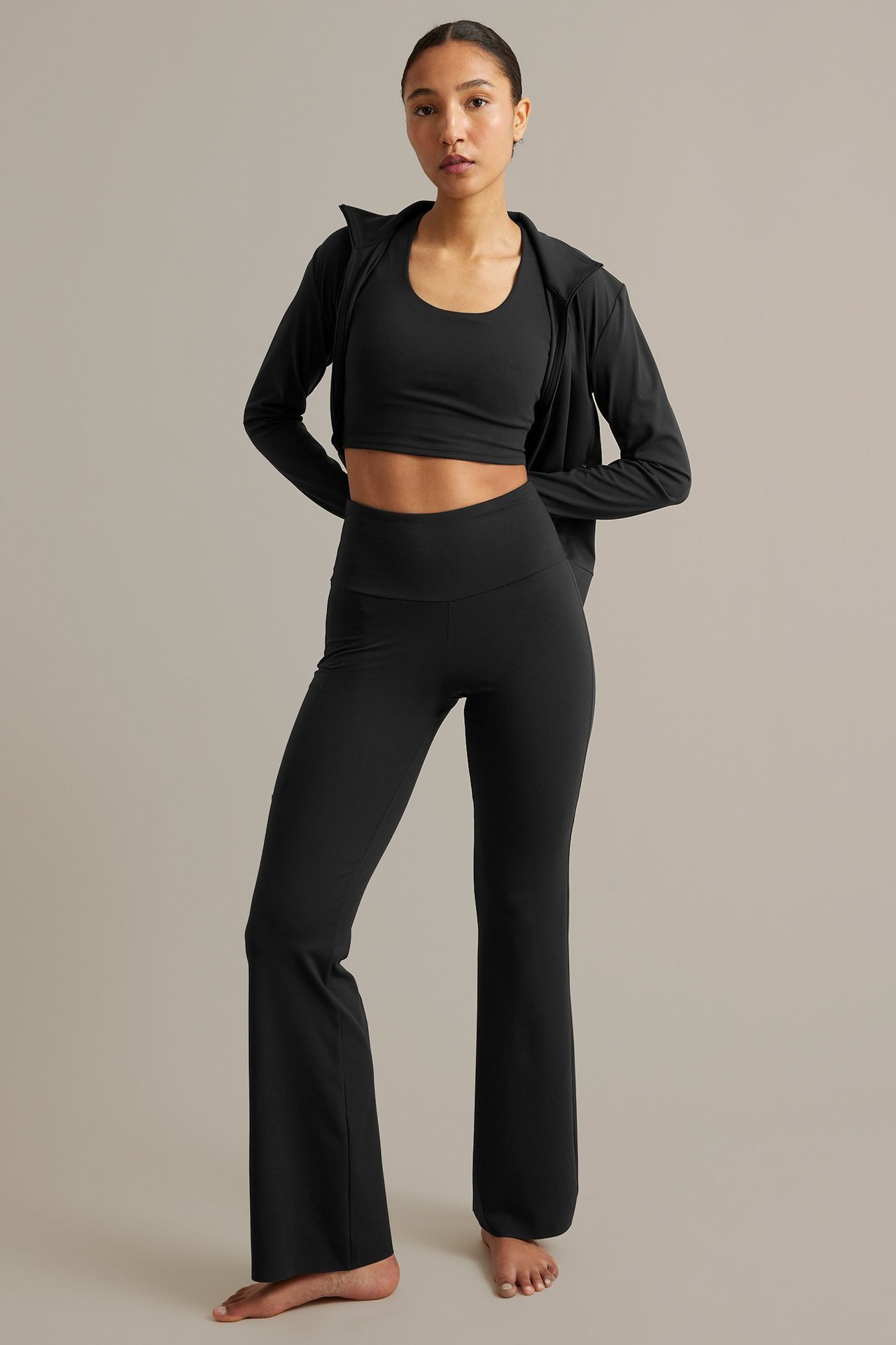 Framifrån, kvinna i svarta höga bootcut träningstights med ficka, sport-BH och träningsjacka med dragkedja.