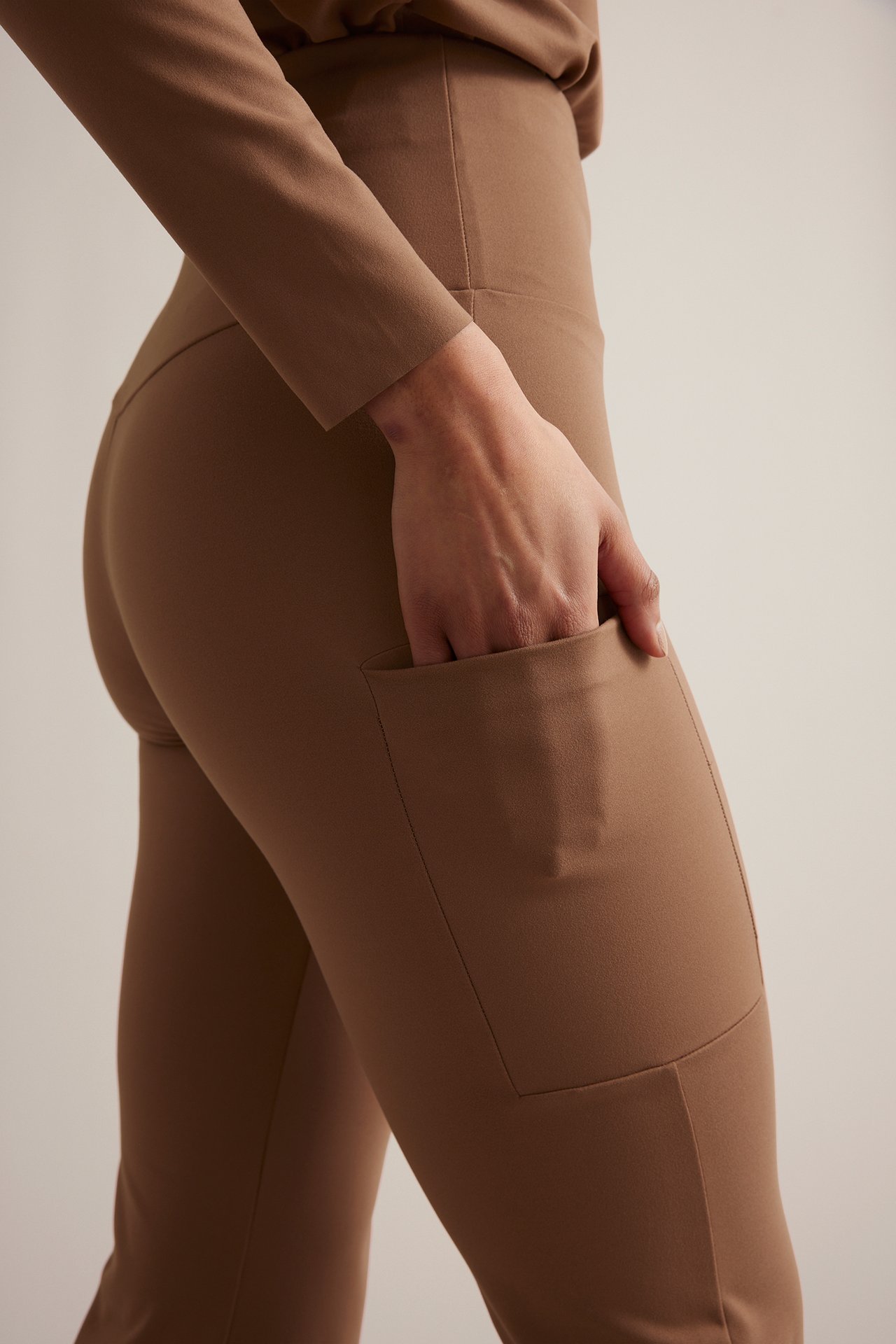 Sidovy av kvinna i bruna högmidjade bootcut träningstights med benficka, med handen i fickan.
