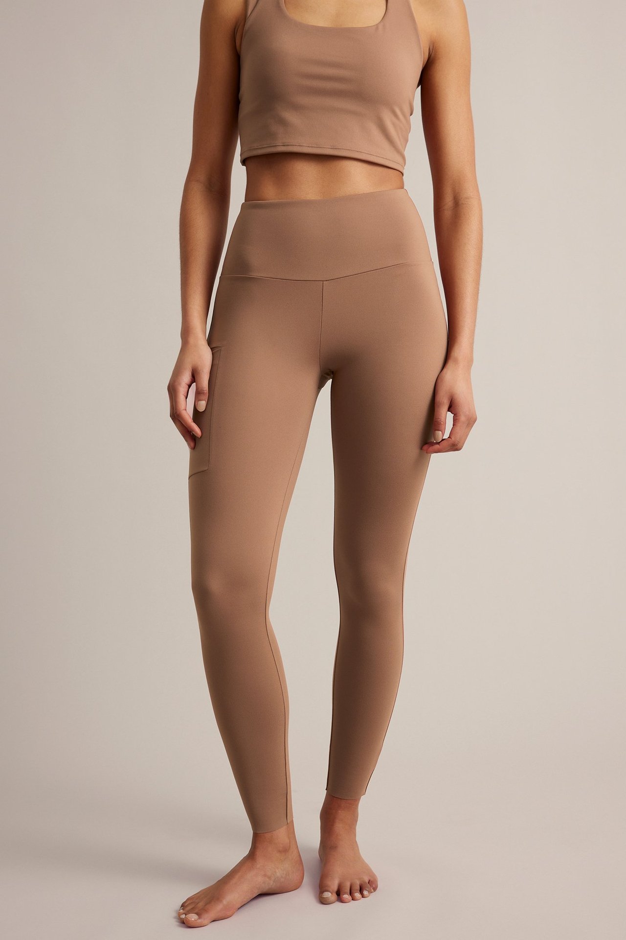 Framifrån, kvinna i bruna träningstights med hög midja, sidoficka och matchande kort topp.