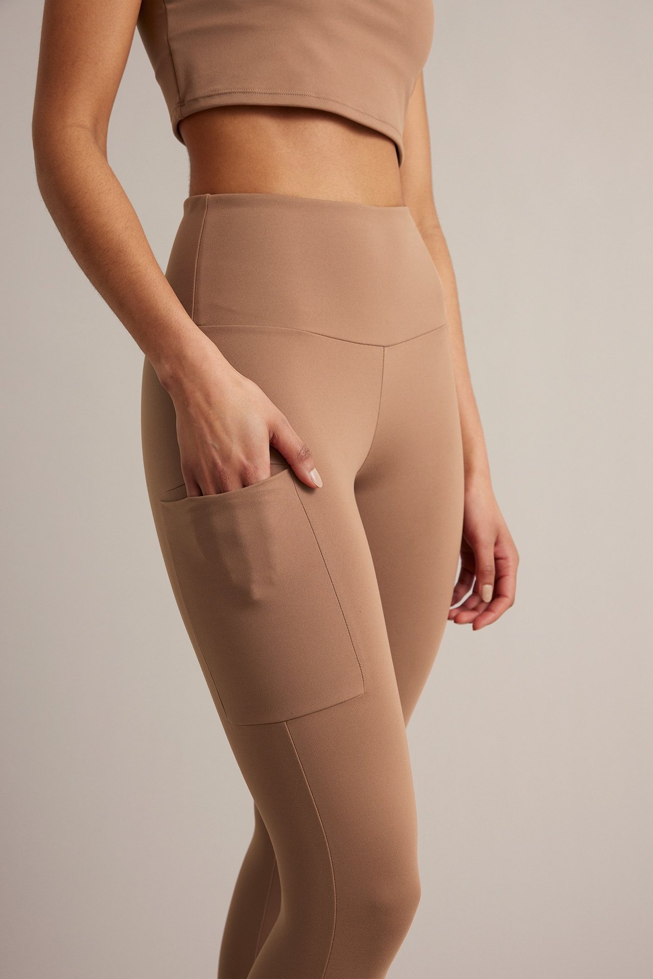Sida vy av kvinna i bruna högmidjade träningstights med sidoficka och matchande kort topp. Hennes hand är i fickan.