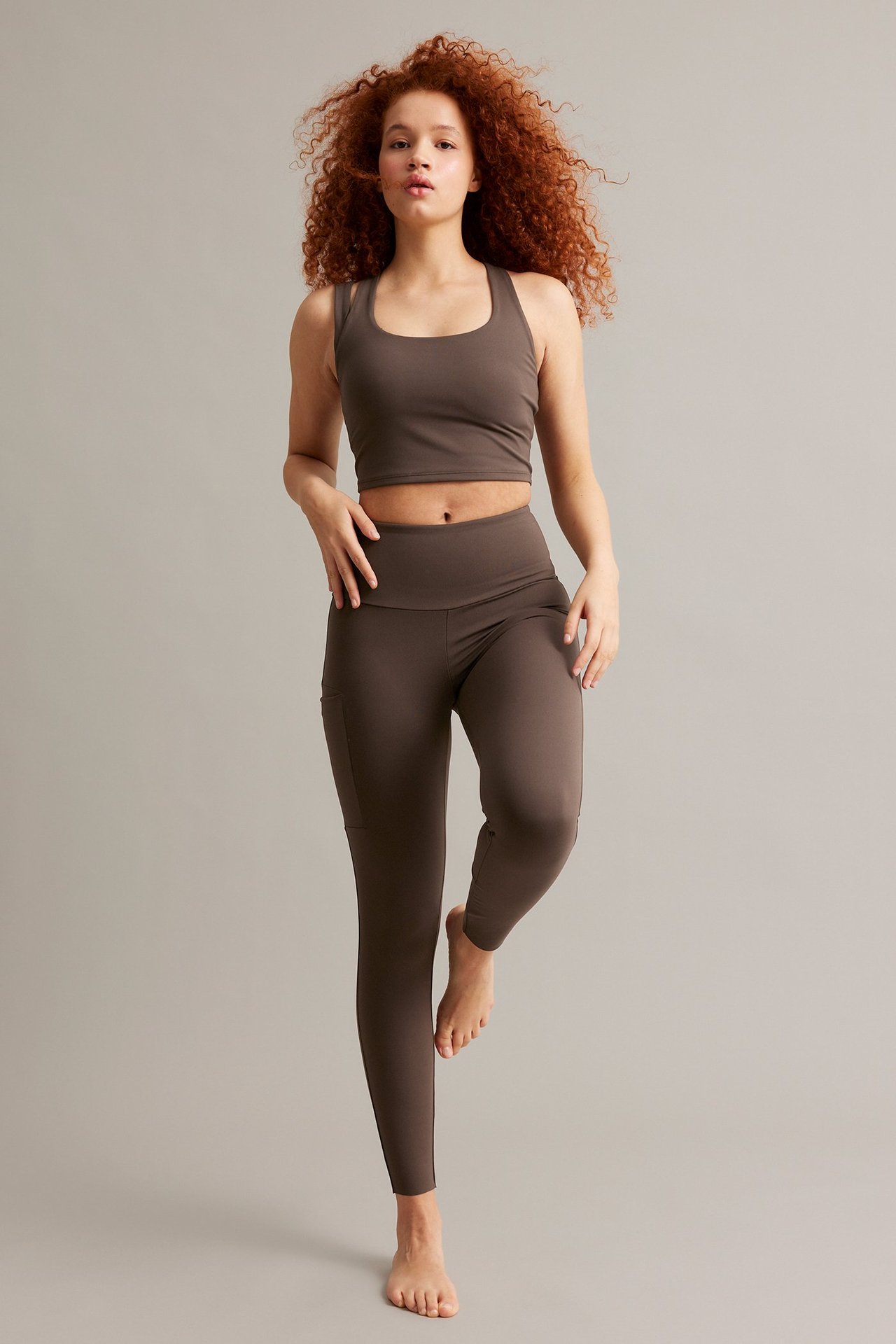 Z przodu: kobieta w brązowych legginsach treningowych z kieszenią i dopasowanym biustonoszu sportowym.
