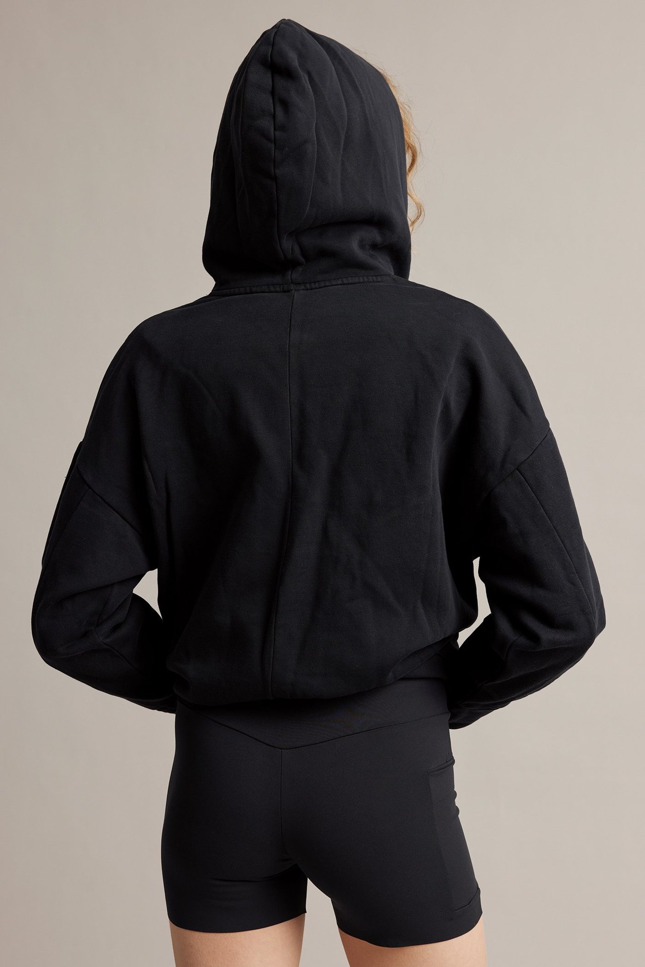 Ryggbilde av kvinne i svarte, høye bikershorts med sidelomme og en svart hettegenser.