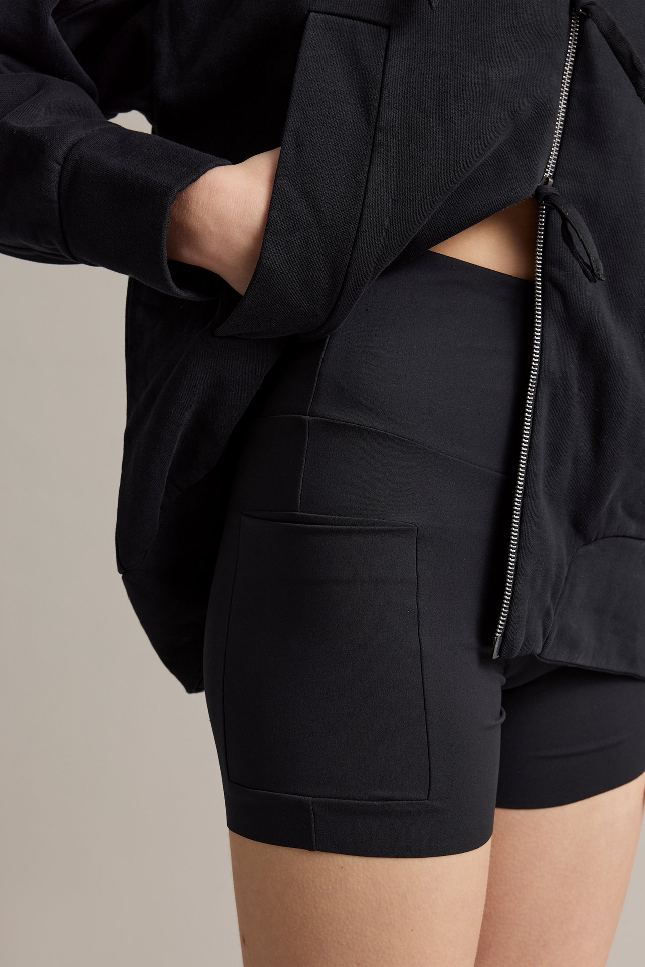 Nærbilde: Kvinne i svarte høywaist bikershorts med sidelomme, under en svart jakke med glidelås.