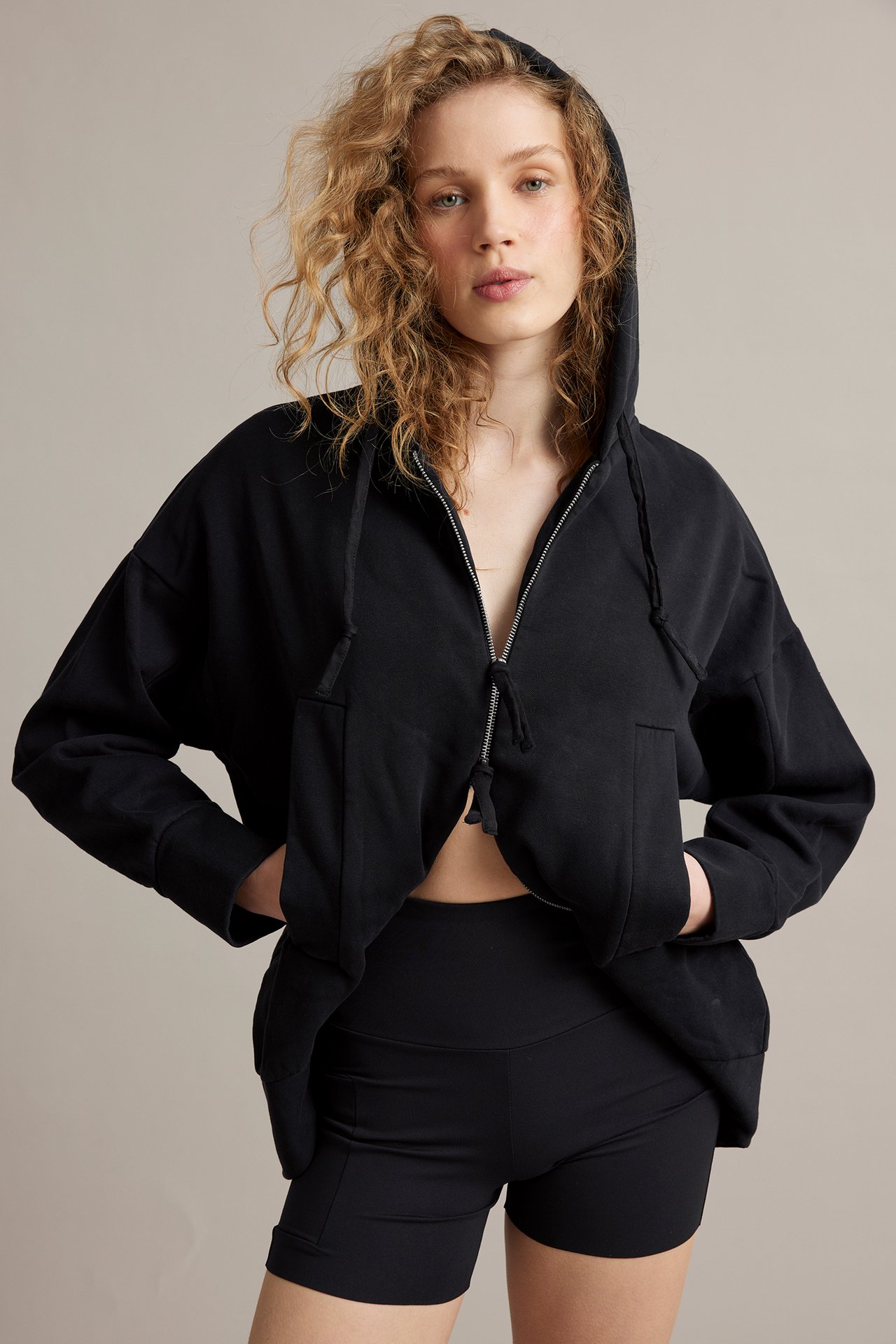 Edestä, nainen mustissa korkeavyötäröisissä pyöräilyshortseissa taskulla, yllään musta huppari.