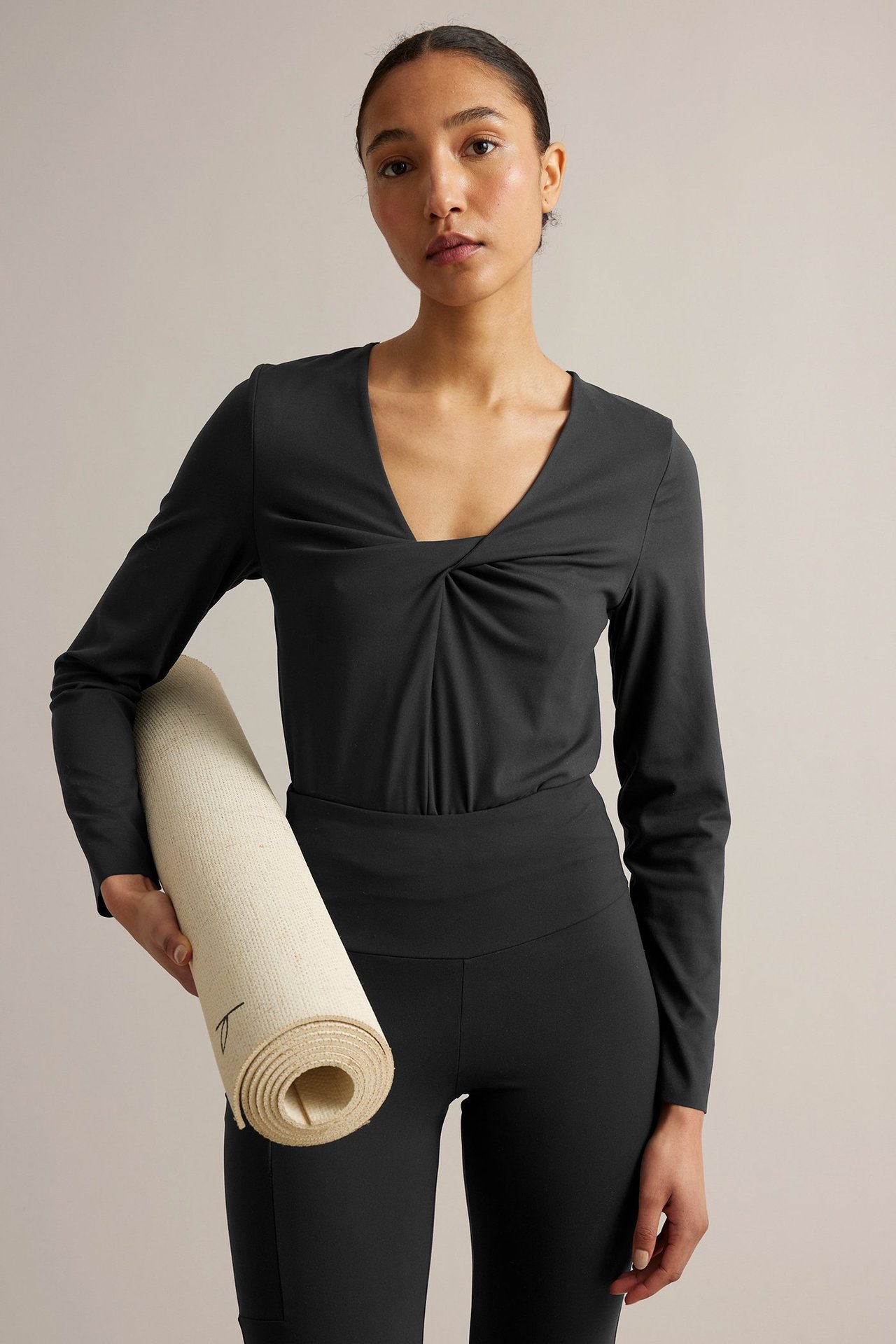 Forfra: Kvinne i svart langermet treningstopp med knutet v-hals og sorte leggings, holder yogamatte.