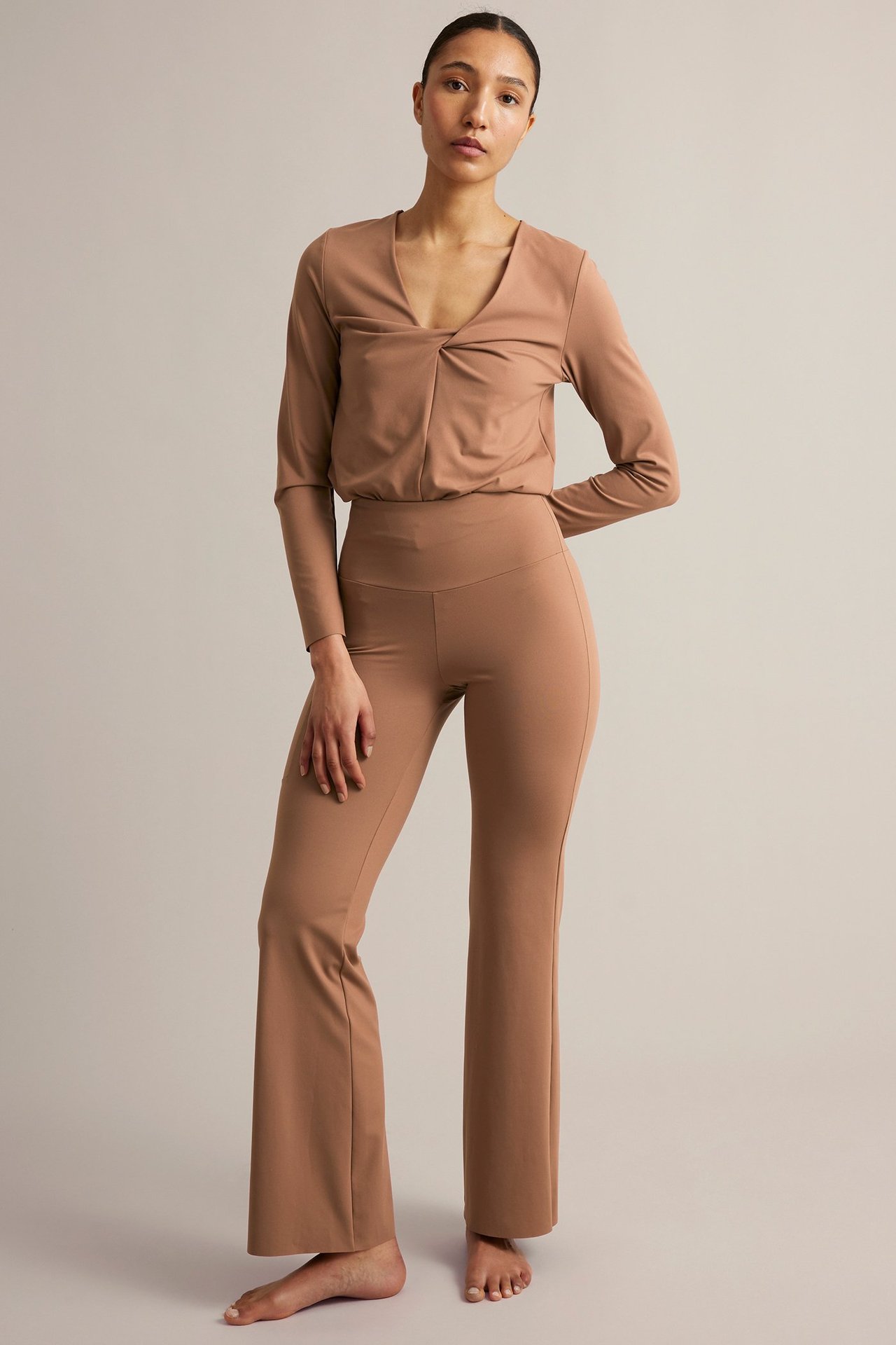 Widok z przodu: Kobieta w brązowym topie treningowym z długim rękawem i wiązaniem V-neck, pasujących spodniach do jogi.