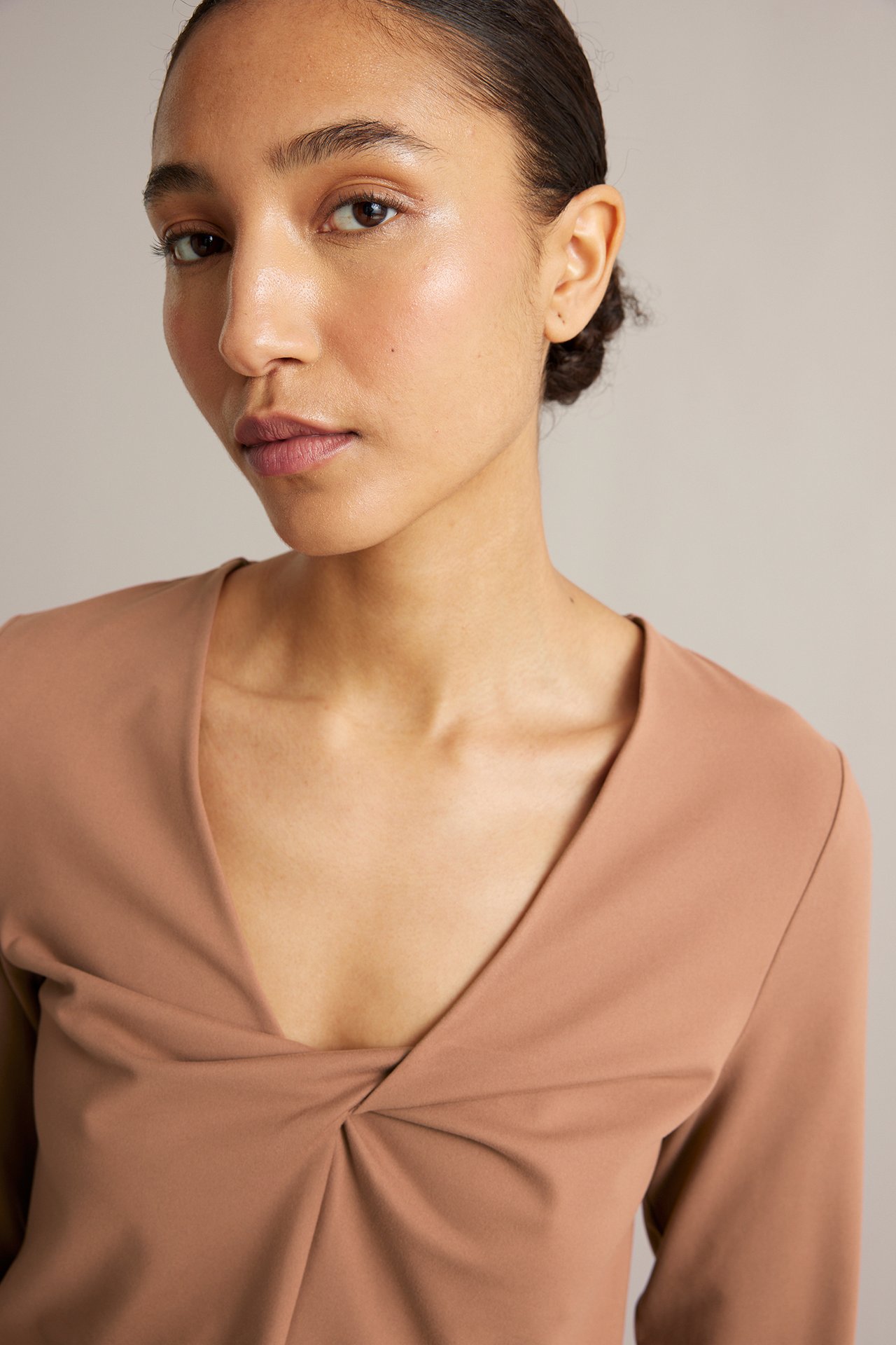 Närbild, framifrån, av en kvinna i ljusbrun träningströja i jersey med dekorativ knut i v-ringningen.