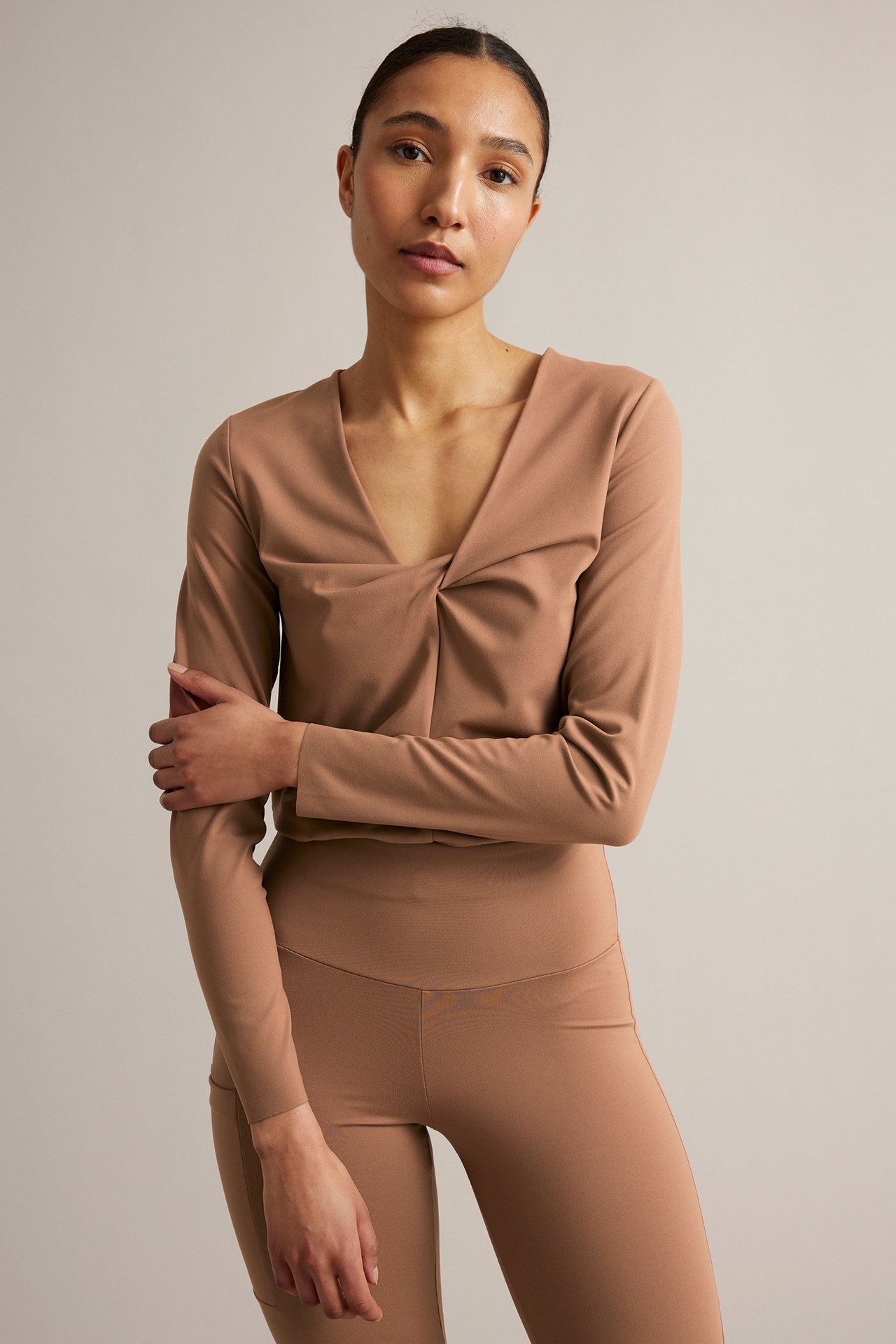 Framsida: kvinna i brun långärmad träningstopp med V-ringning och knut, samt matchande bruna tights.