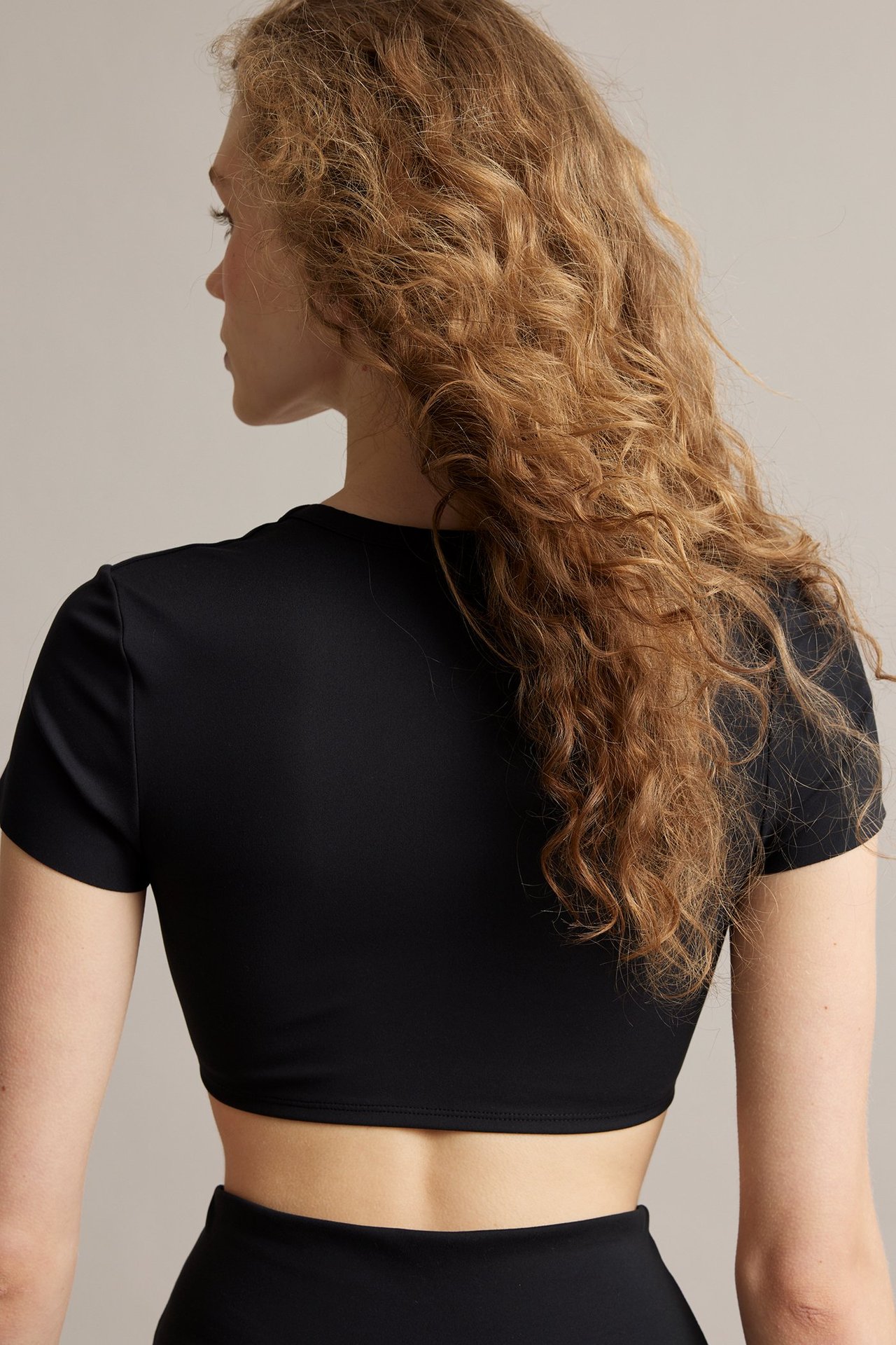Widok z tyłu kobiety w czarnym, dopasowanym, krótko-rękawowym topie crop z elastycznego dżerseju i pasujących dołów.