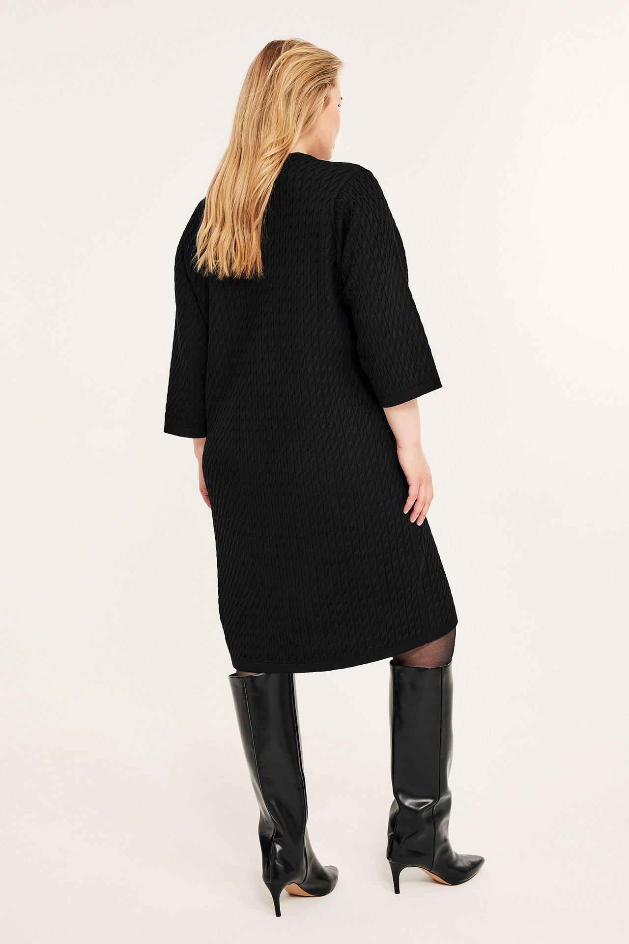 Bakfra av kvinne i svart strukturert kabelstrikket kjole med 3/4-ermer og svarte knelange støvler.