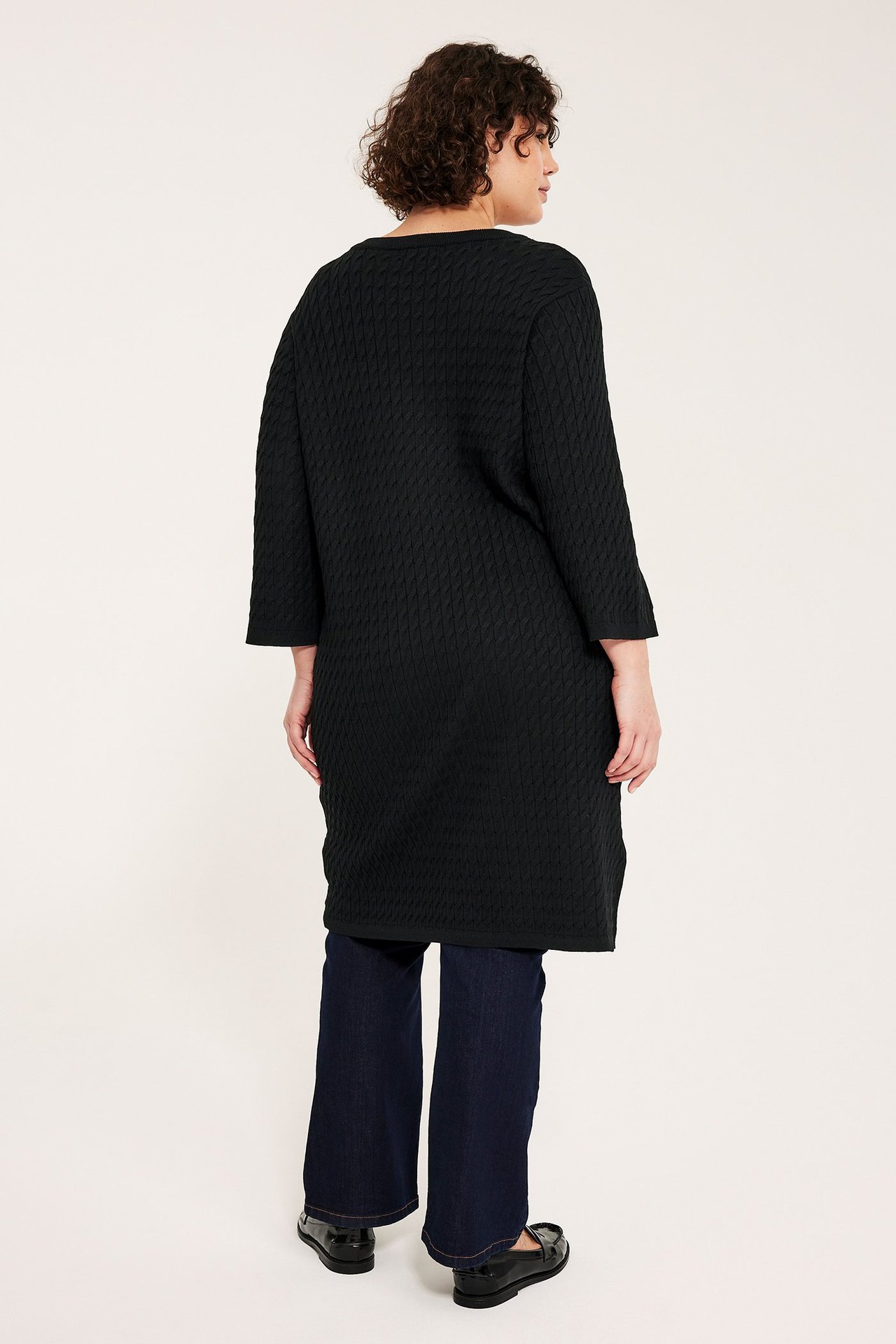 Sidevisning bakfra av kvinne i sort kabelstrikket kjole, mørkeblå jeans og sorte loafers.
