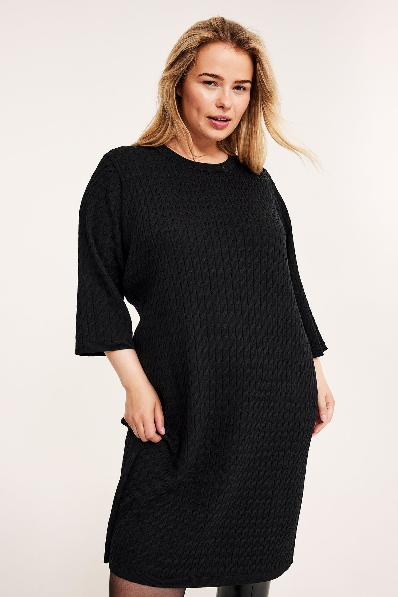 Frontvisning av en kvinne i svart kabelstrikket knelang kjole med 3/4-ermer og rund hals.