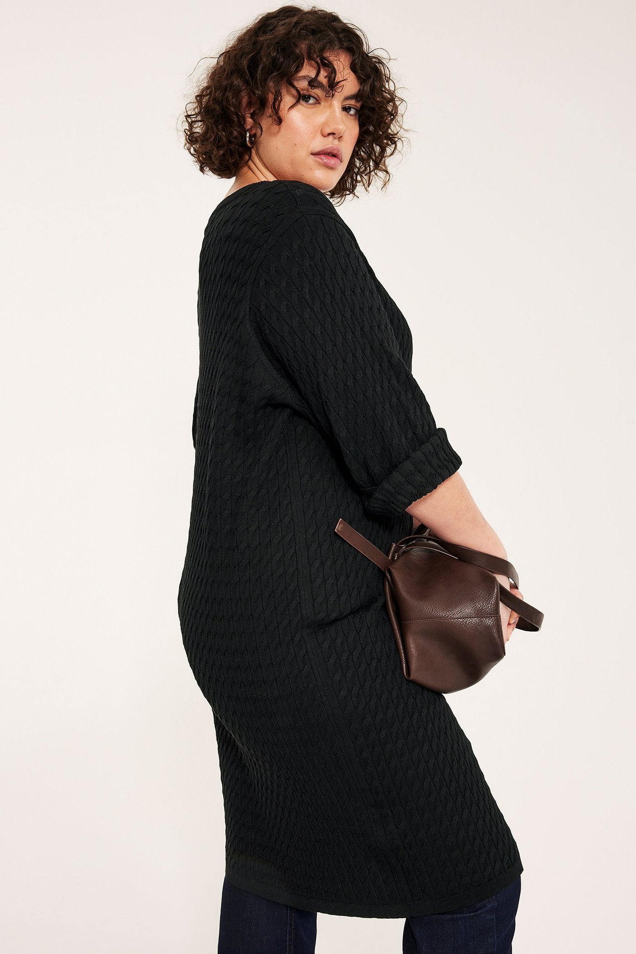 Sivukuva: Nainen mustassa palmikkoneuleisessa midimekossa, kääritetyt hihat, tummat farkut ja ruskea vyölaukku.