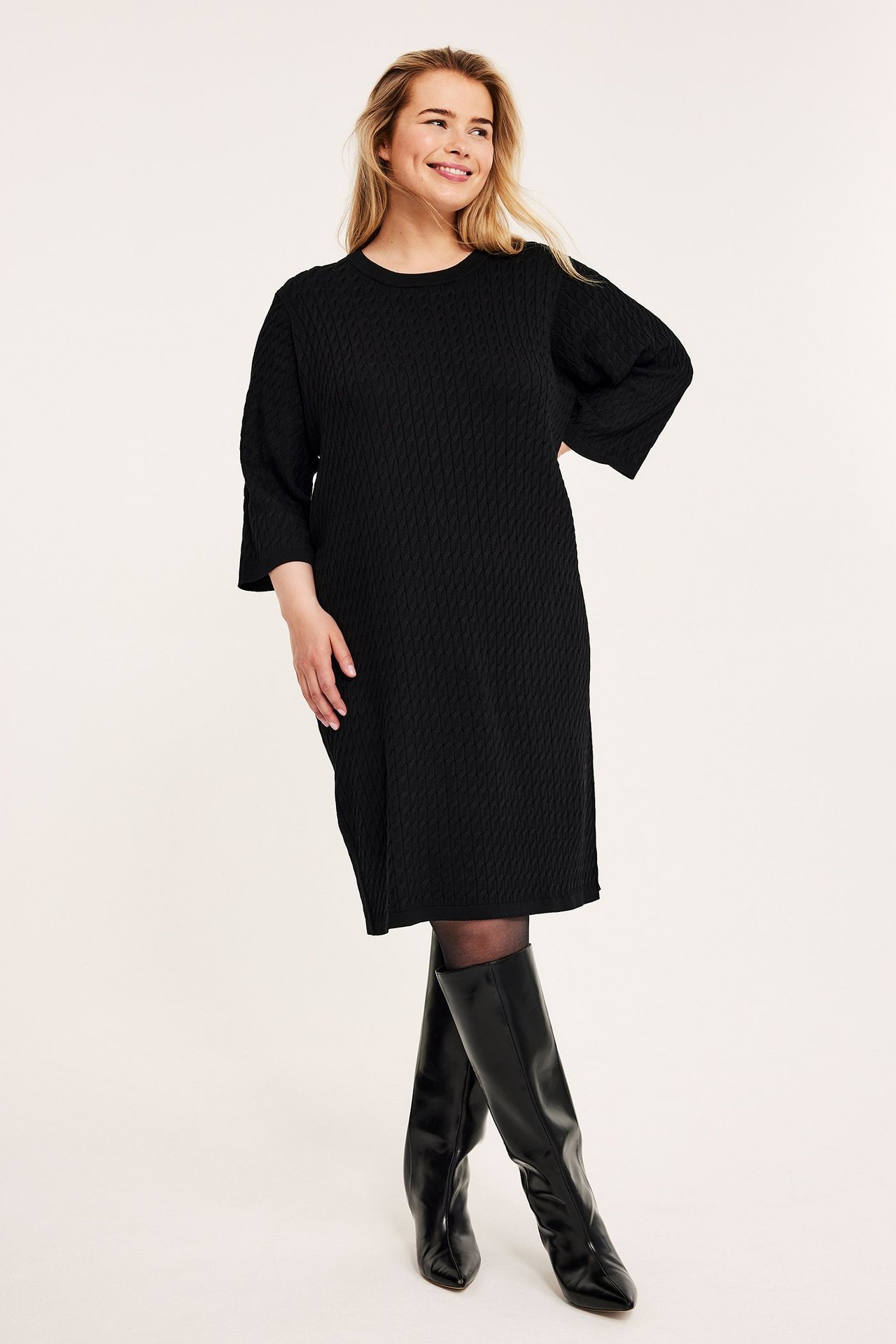 Forfra, kvinne i svart strukturert kabelstrikket kjole, svarte strømpebukser og knelange støvler.